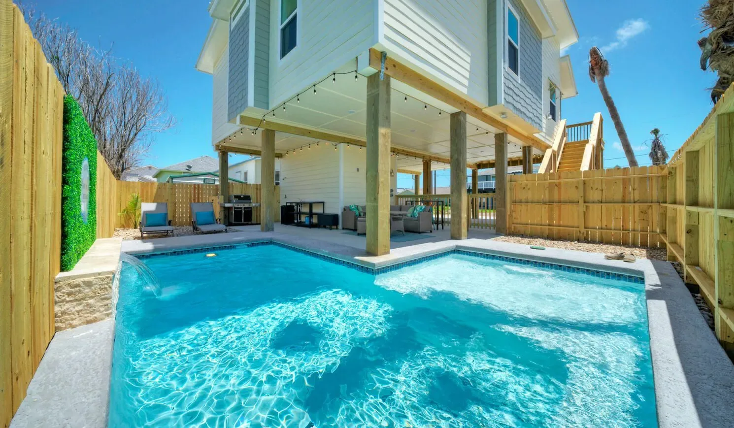 A swimming pool sits beside a raised house and wooden fencing.