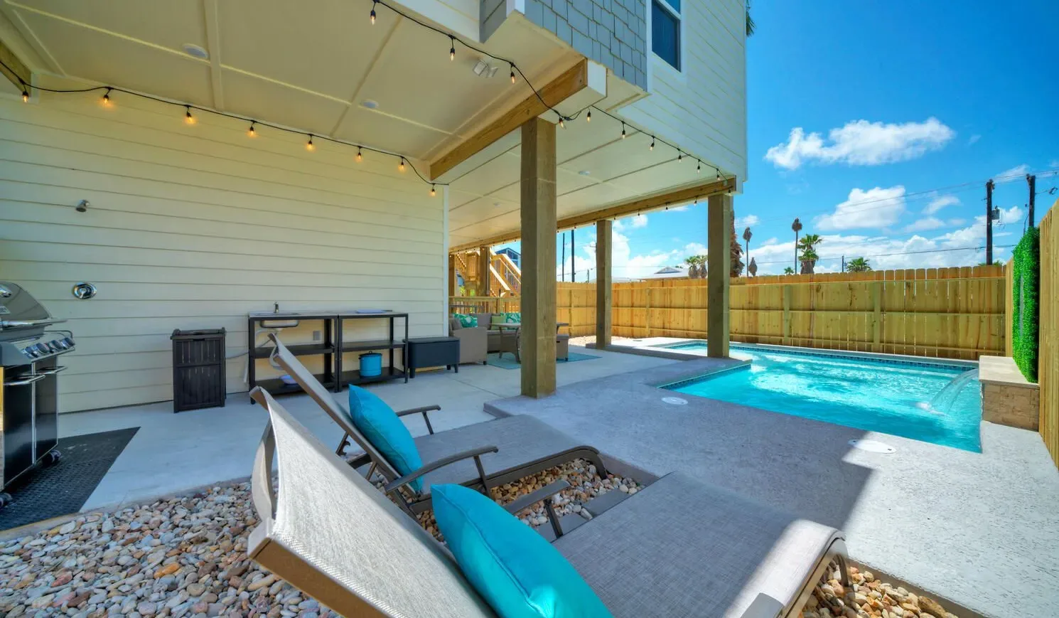 An outdoor pool sits beside a covered patio with seating and string lights.