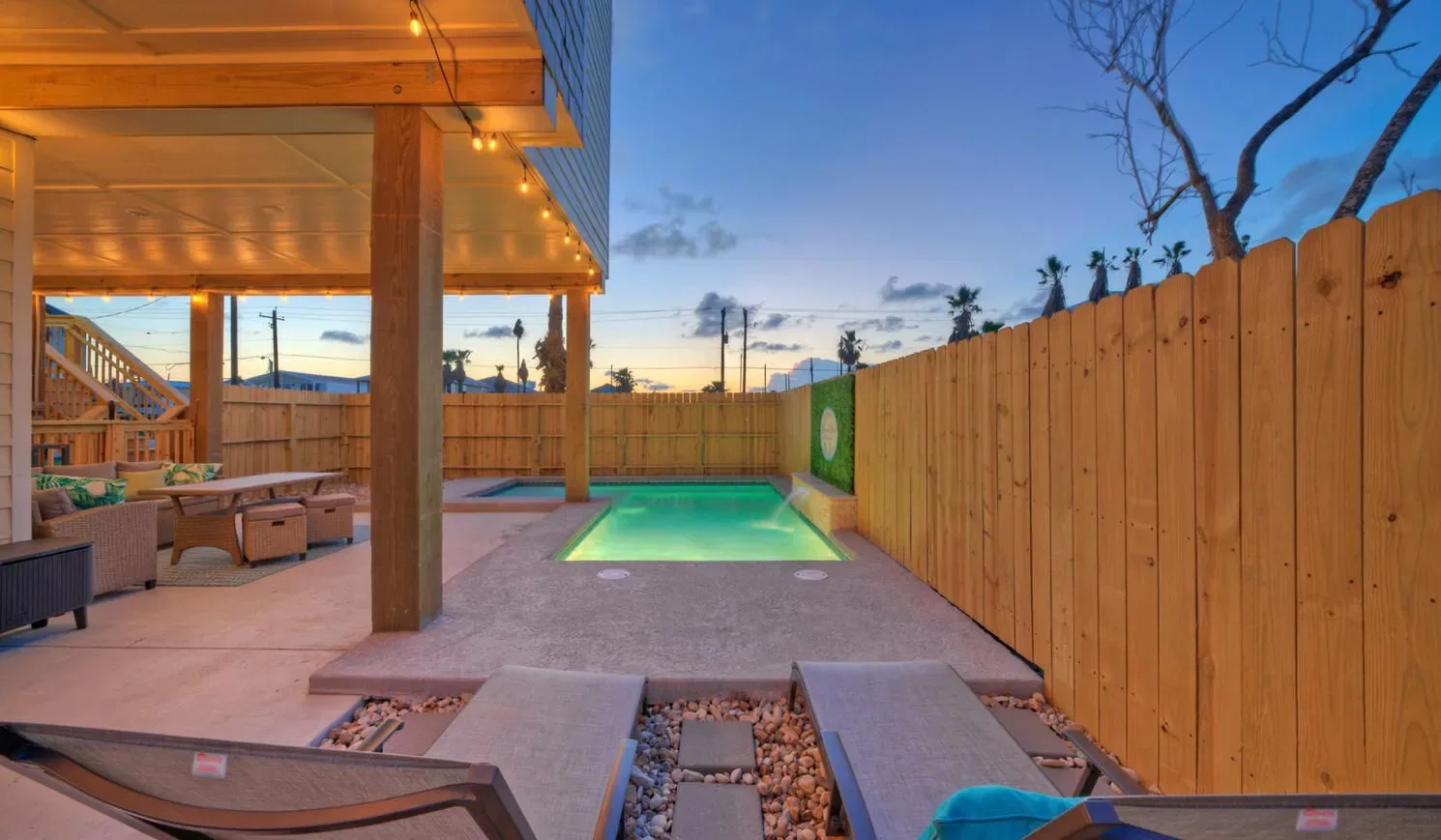An outdoor patio with a lit pool, lounge seating, and a wooden fence at dusk.