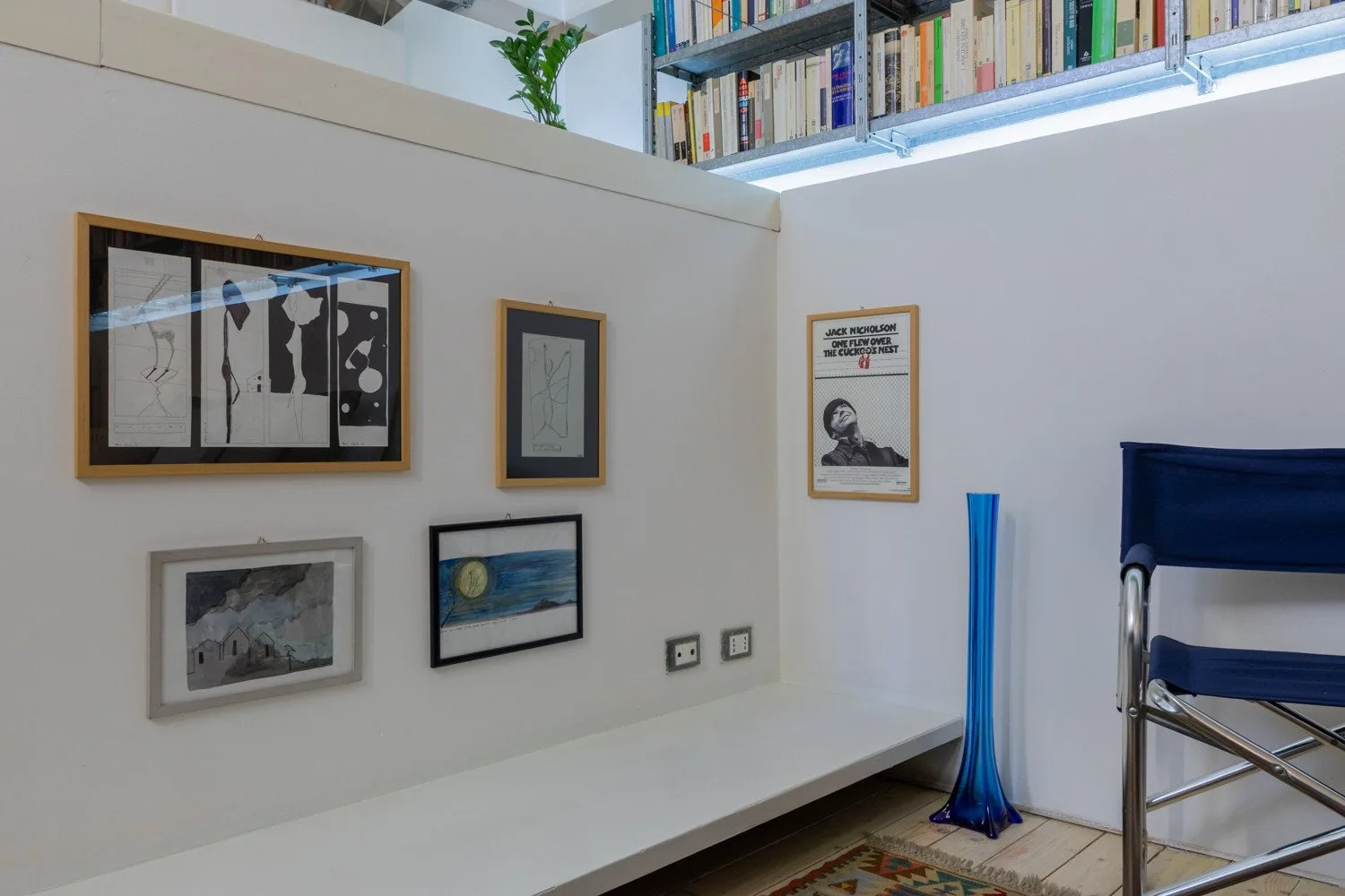 Una sala luminosa con obras enmarcadas y libros en un estante alto.