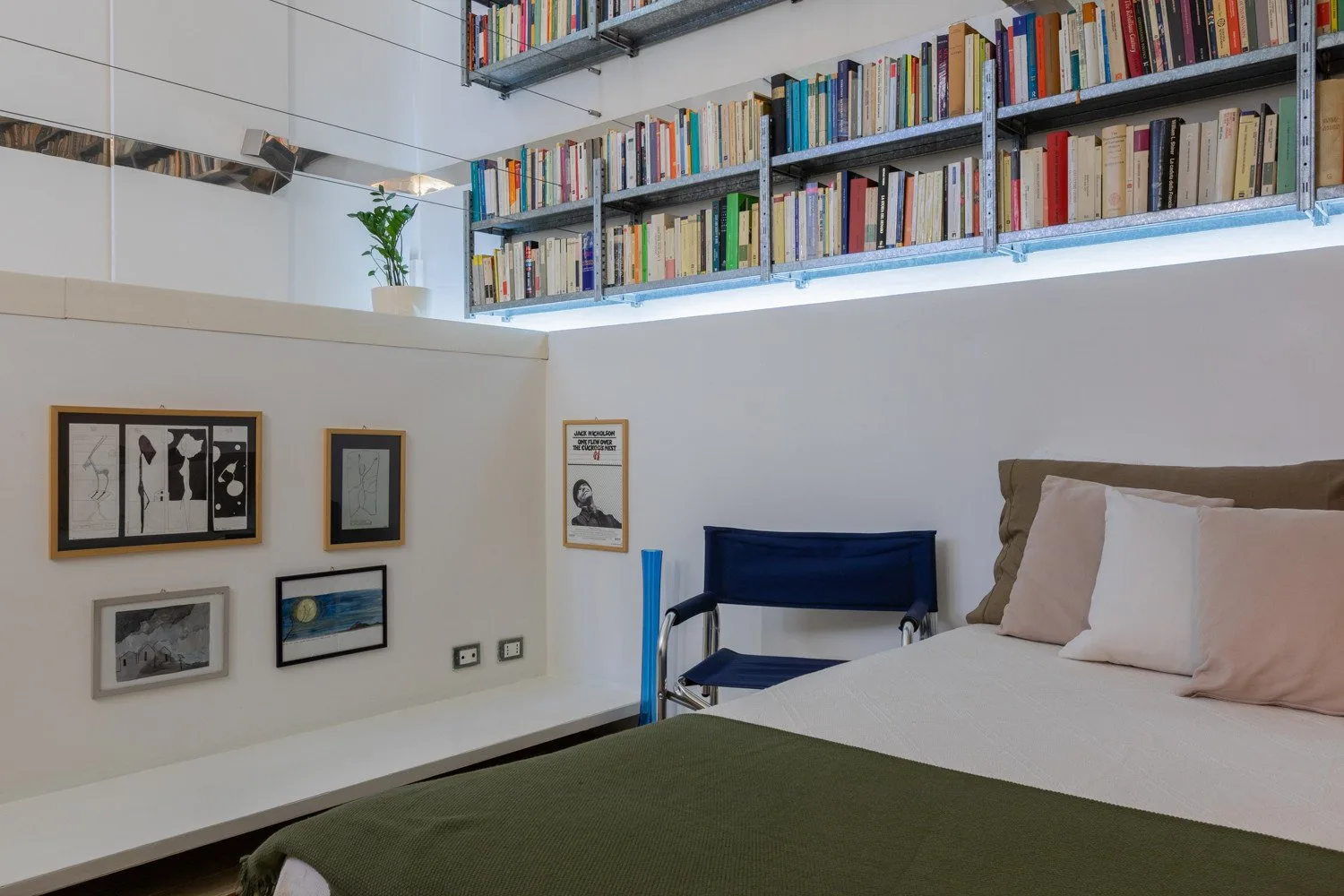 Un dormitorio con una cama, una silla azul y estanterías de libros en la pared superior.