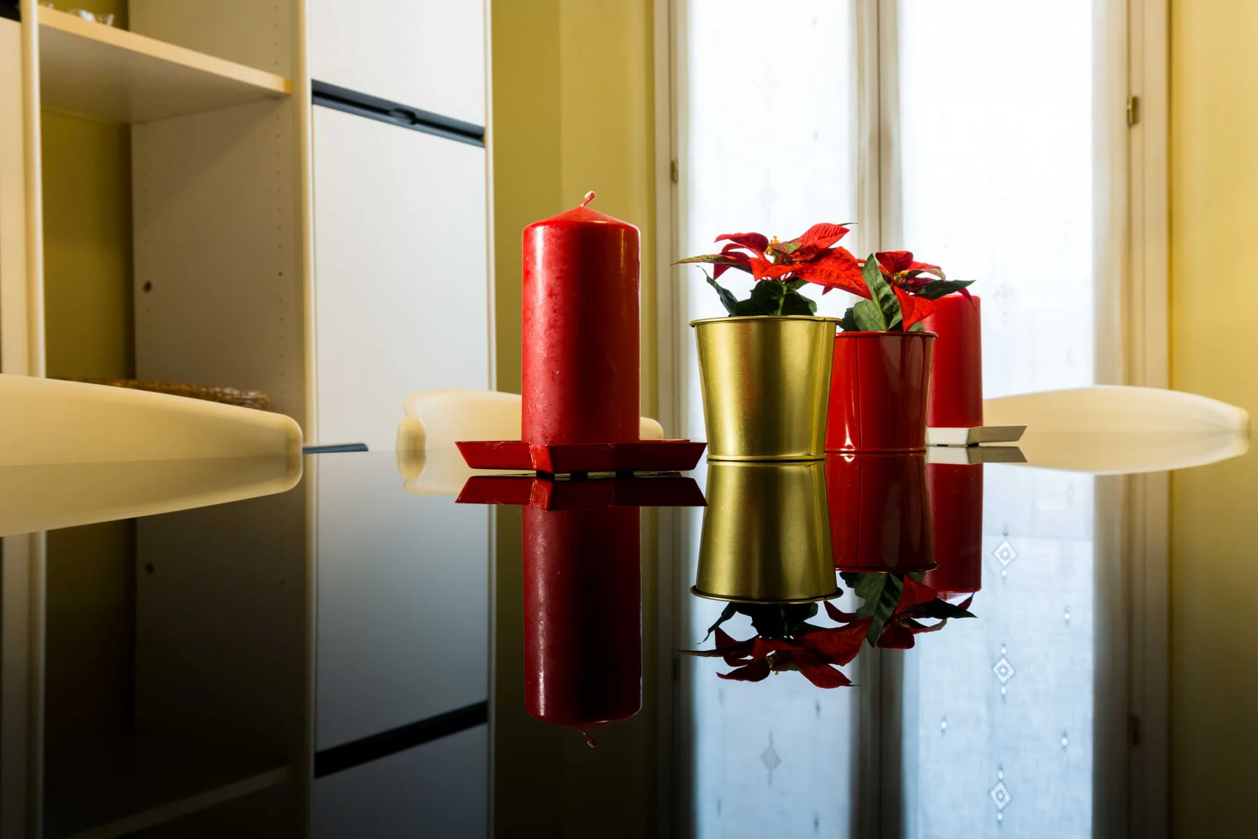A glass dining table holds a red candle and potted poinsettias beside a bright window.