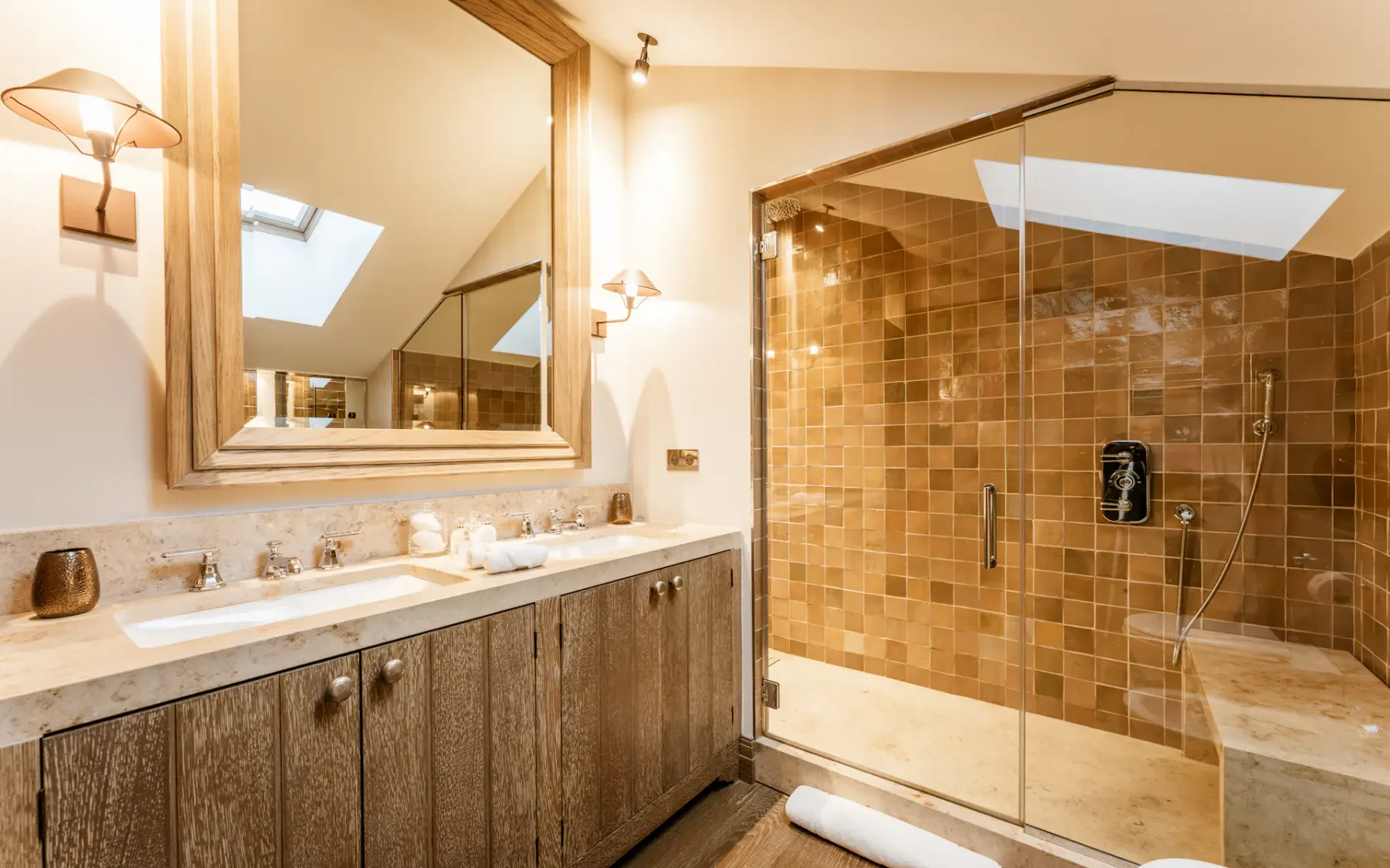 Baño moderno con armarios de madera, dos lavabos, un gran espejo y una ducha con mampara de cristal y azulejos marrones. Iluminación suave y una claraboya.