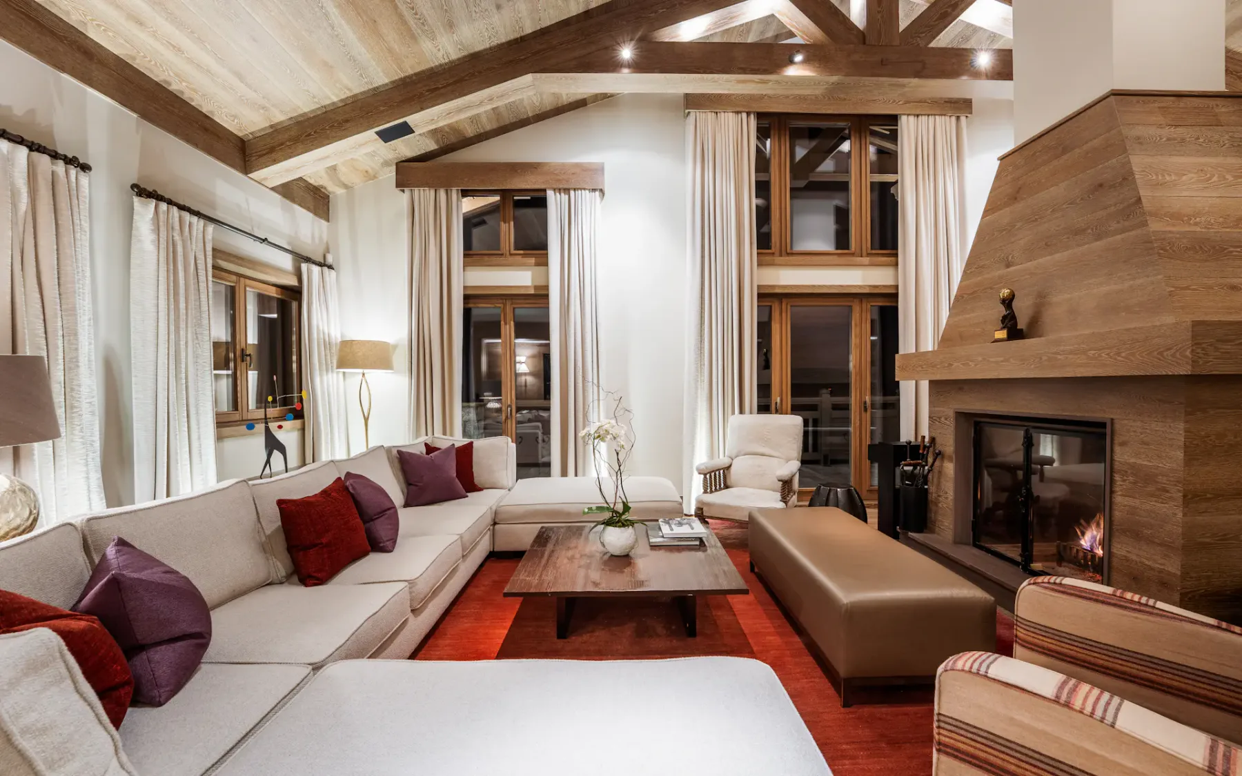 Acogedora sala de estar con sofás beige, cojines rojos, mesa de centro de madera, chimenea y grandes ventanas con cortinas. Iluminación cálida y vigas de madera.