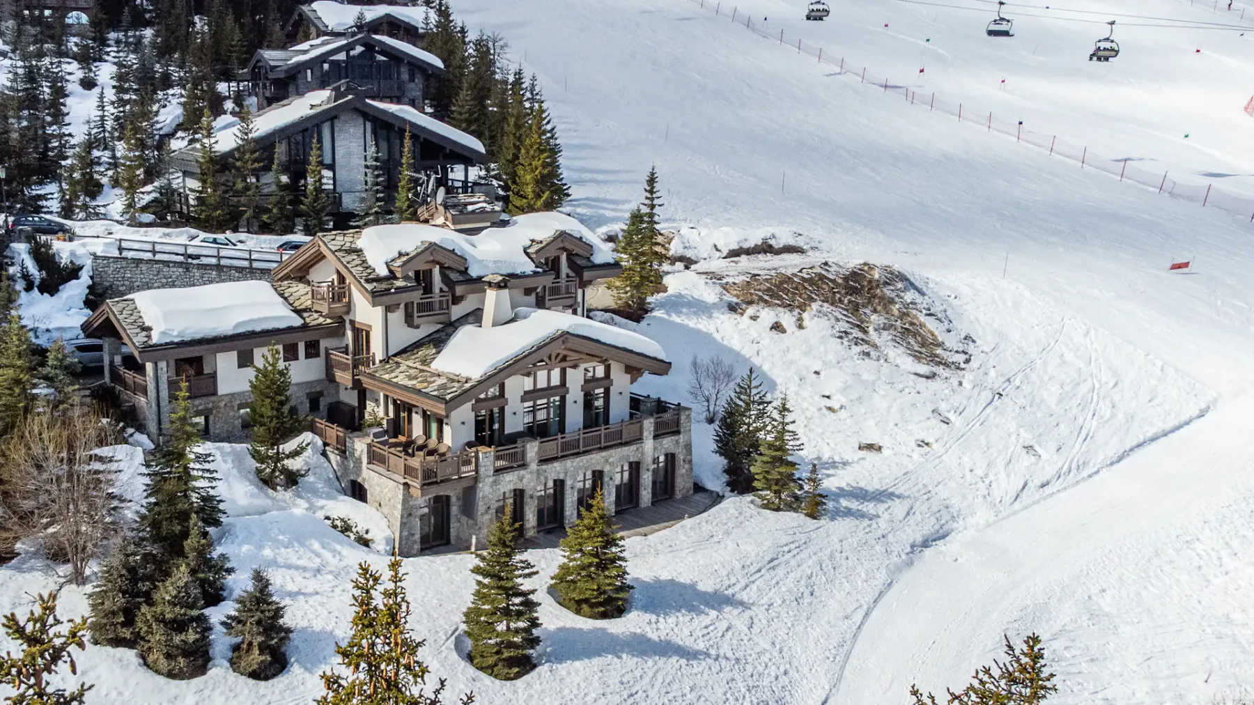 Chalet de montaña rodeado de nieve, cerca de una pista de esquí con telesillas al fondo y pinos dispersos alrededor.