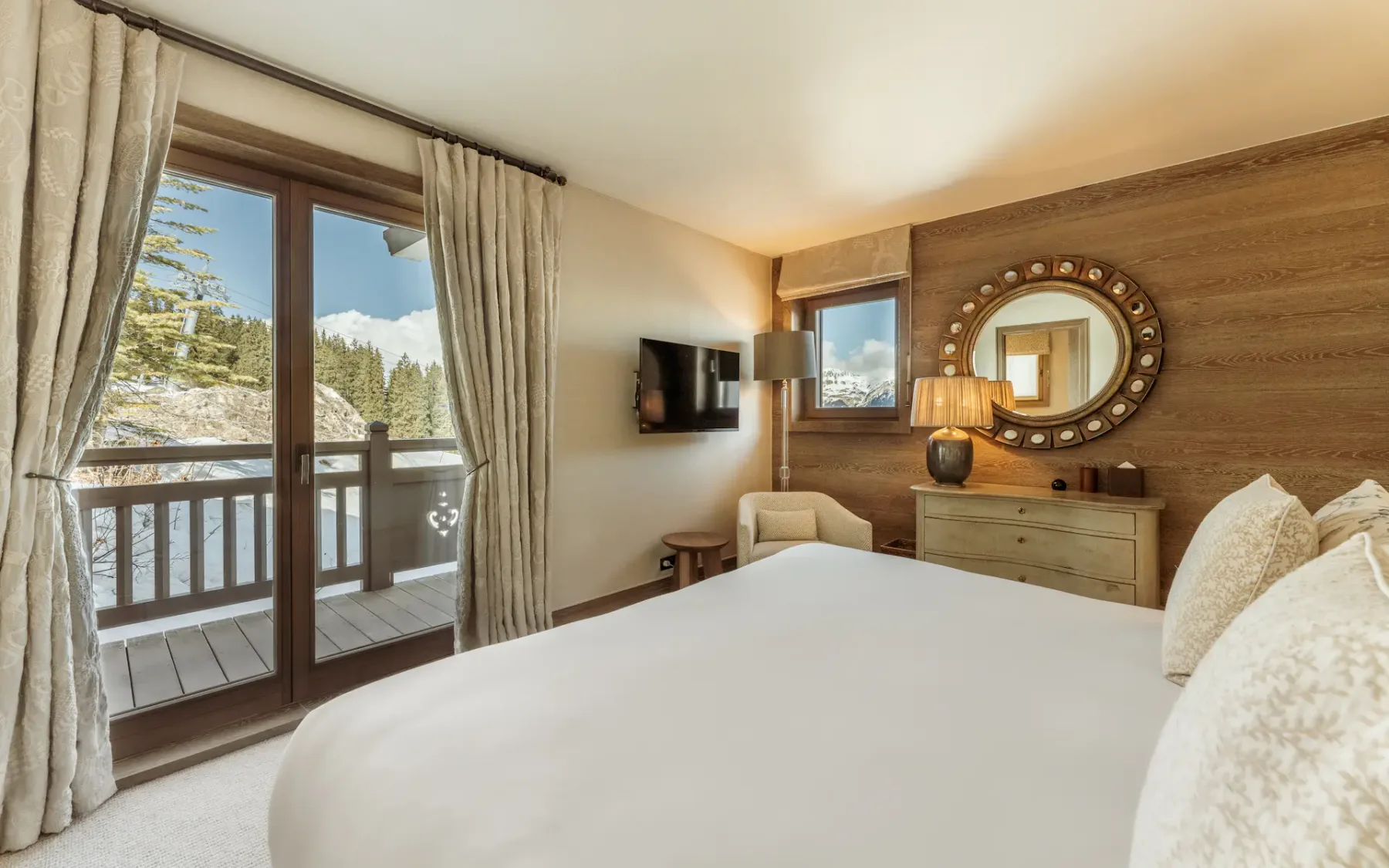 Acogedor dormitorio con una cama grande, detalles de madera, un espejo redondo y vistas a montañas nevadas a través de puertas de cristal que dan a un balcón.