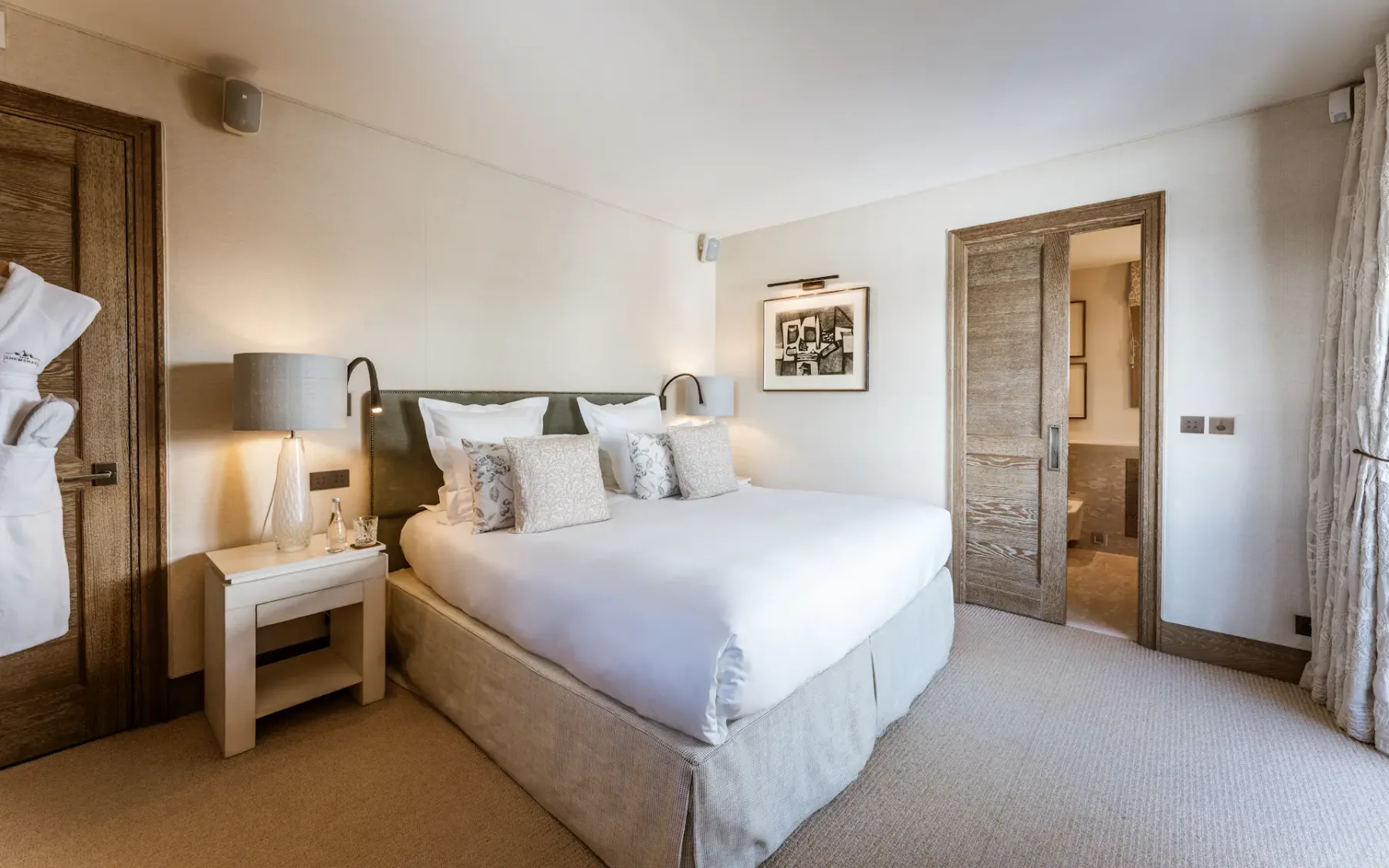 Acogedora habitación de hotel con una cama grande, dos mesitas de noche con lámparas, un albornoz colgado en la pared y una puerta que da a un baño.
