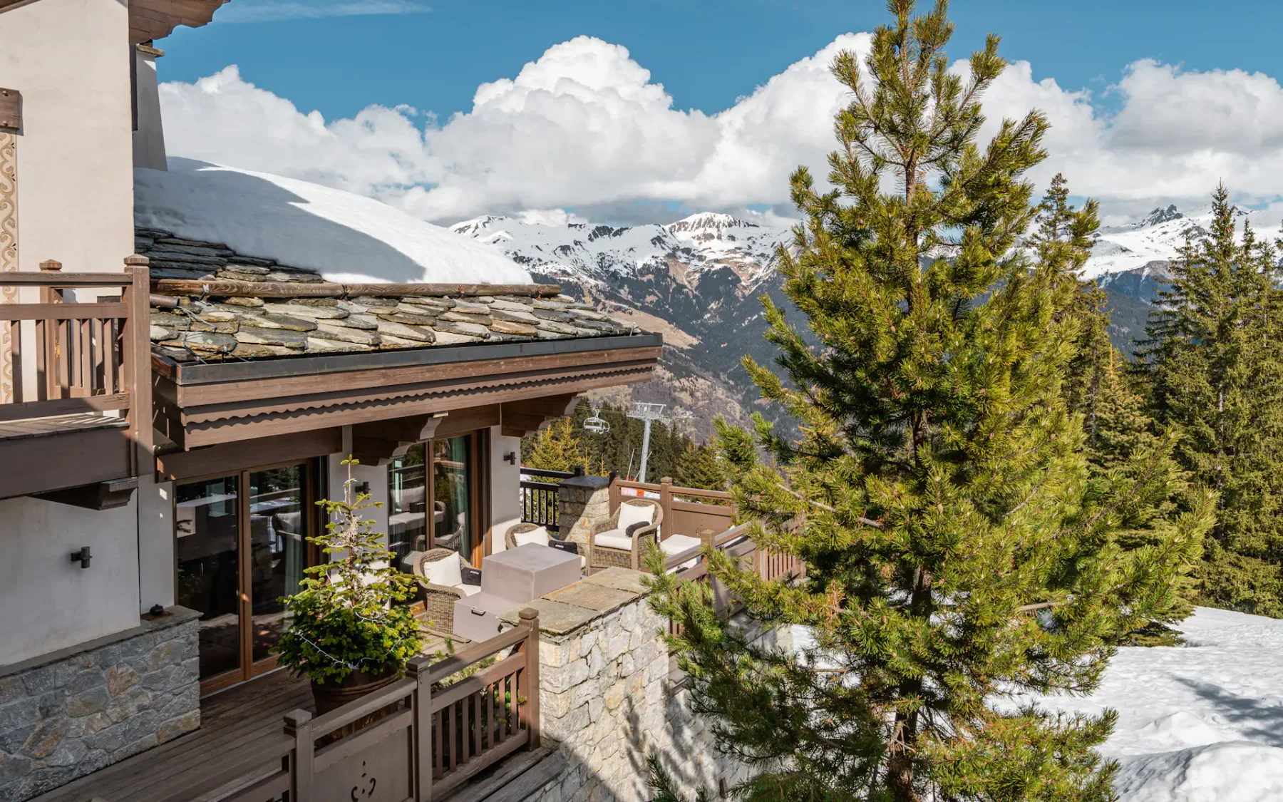 Chalet de montaña con una terraza de piedra, rodeado de picos nevados y pinos bajo un cielo azul con nubes esponjosas.