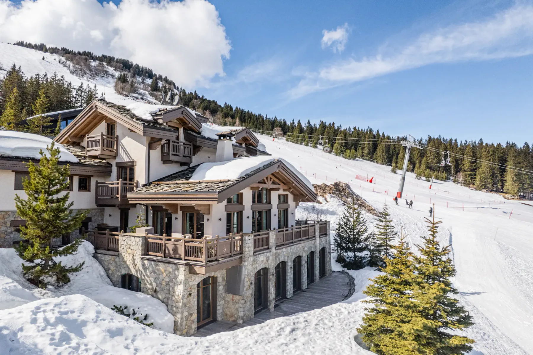 Un gran chalet con balcones de madera y detalles de piedra, rodeado de nieve y pinos, cerca de una pista de esquí bajo un cielo azul despejado.