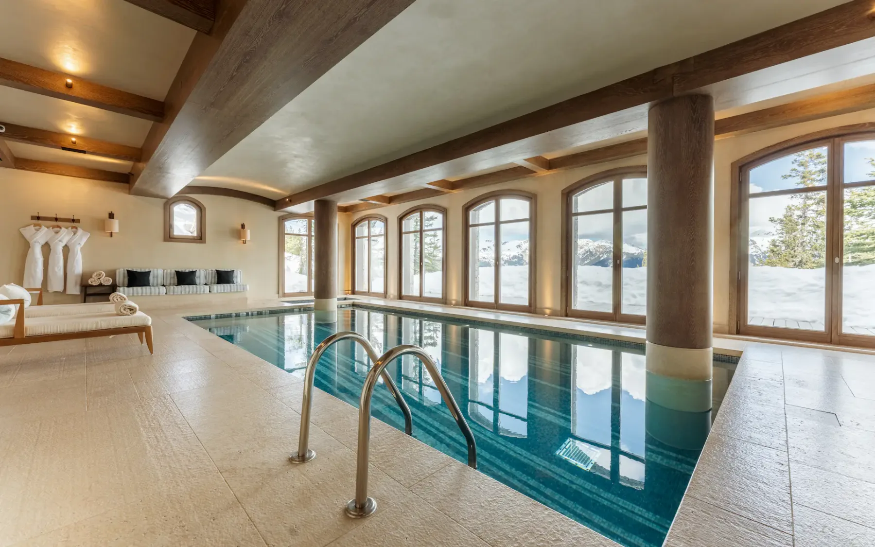 Piscina cubierta con grandes ventanas arqueadas, tumbonas y vigas de madera, con vistas a un paisaje nevado.