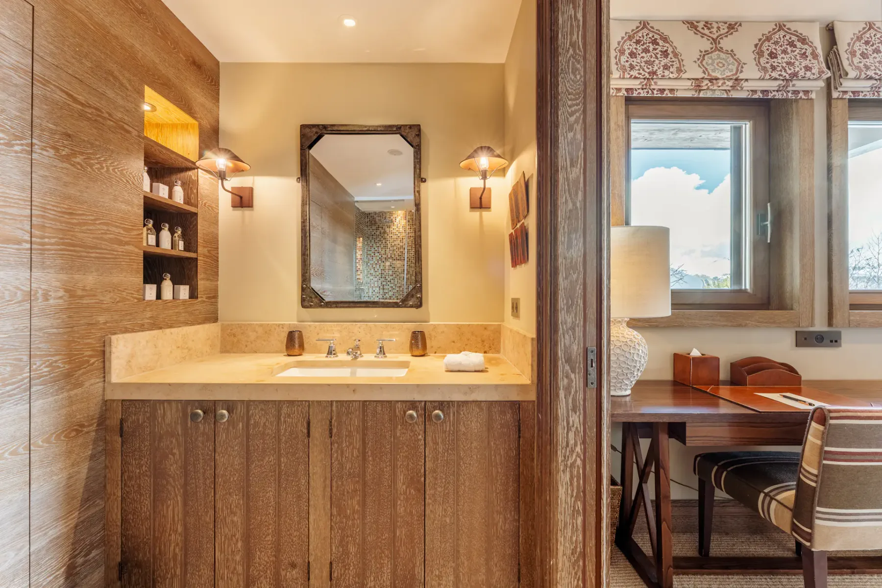 Acogedor baño con armarios de madera, un espejo y apliques de pared. Escritorio contiguo con una lámpara y vistas al cielo desde la ventana.