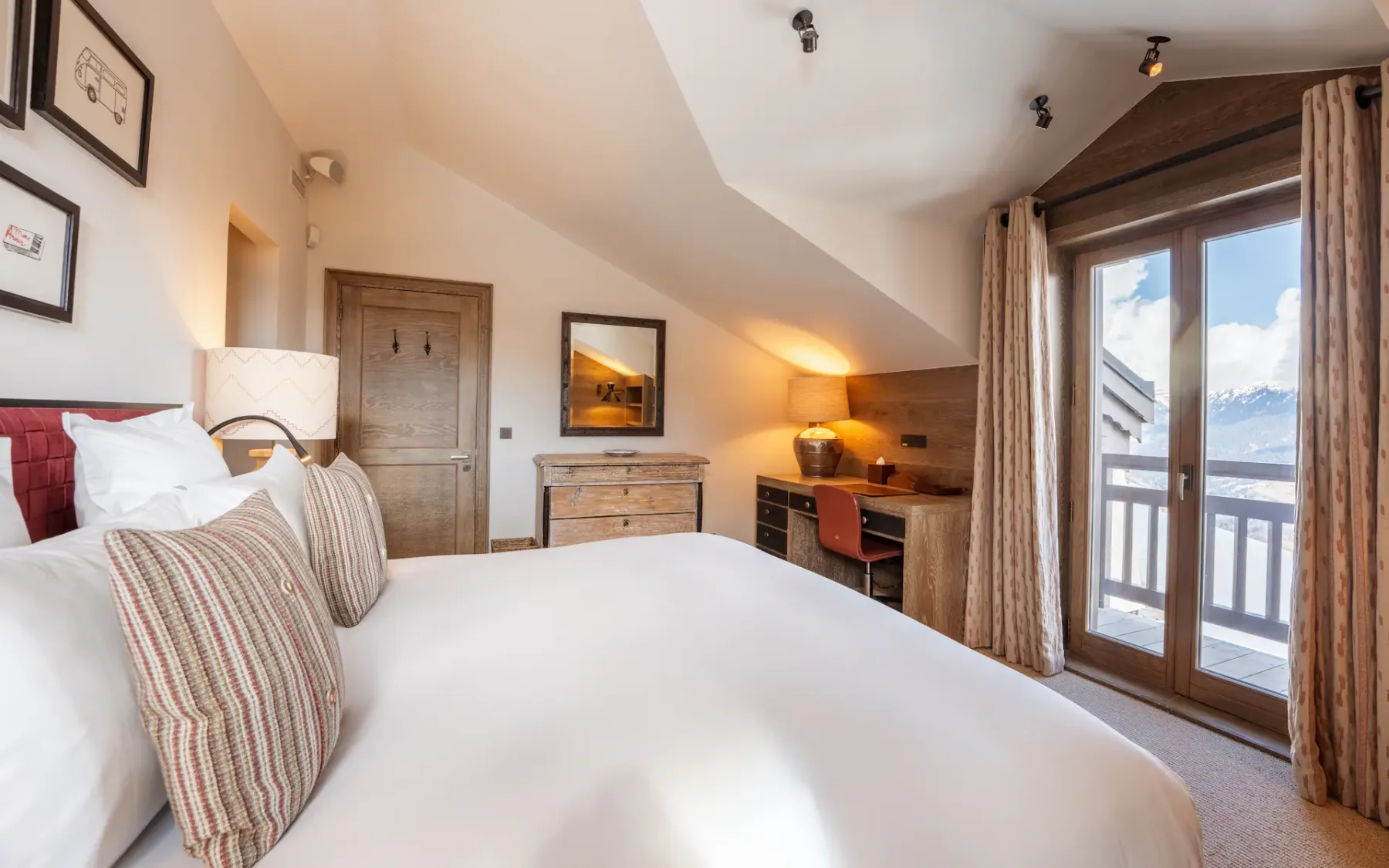 Acogedor dormitorio con una cama grande, muebles de madera, un escritorio y una puerta al balcón con vistas a la montaña. Iluminación cálida y tonos neutros.