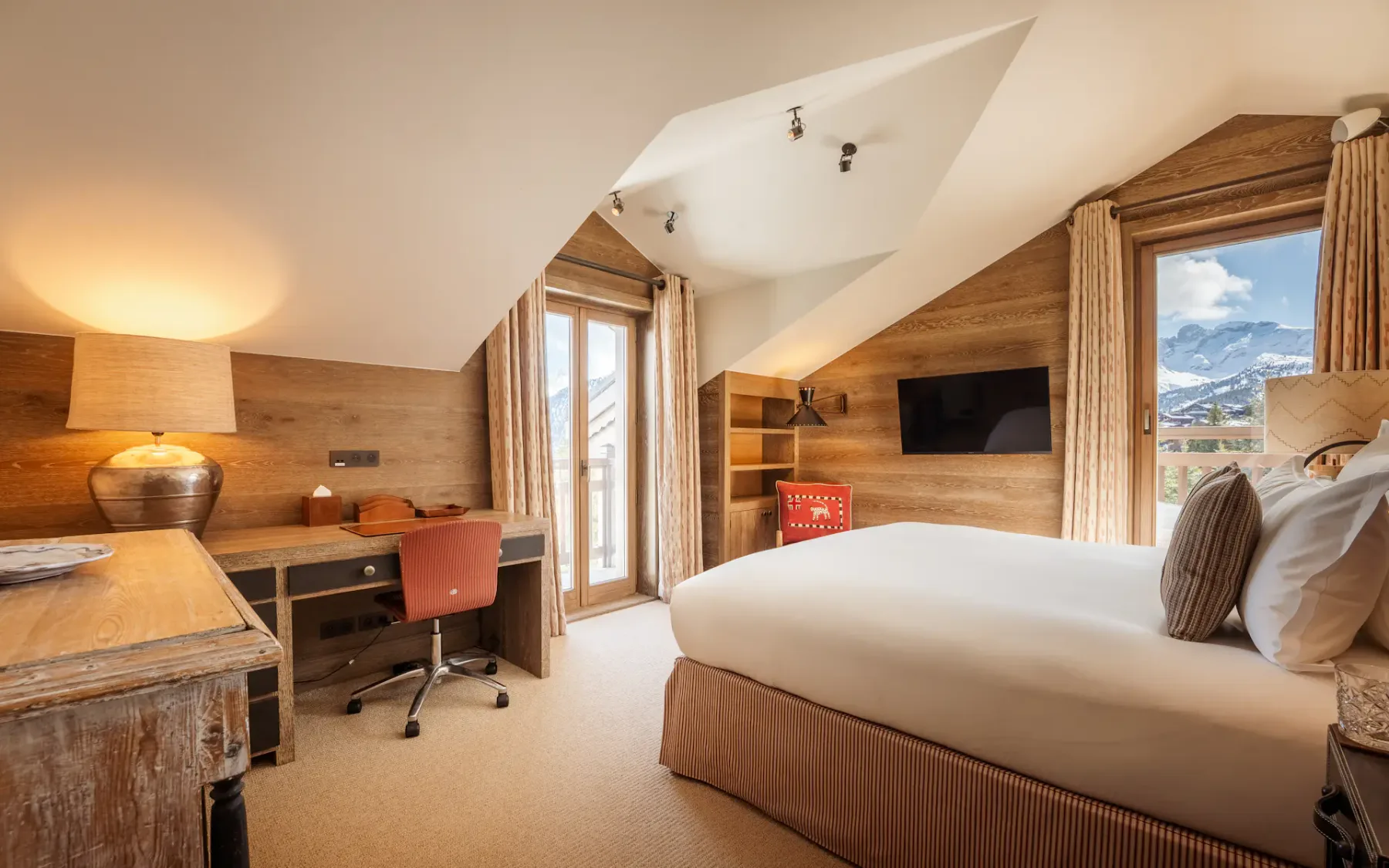 Acogedor dormitorio con techo inclinado, paredes de madera, cama, escritorio, silla y una ventana con vistas a la montaña. La iluminación cálida crea un ambiente acogedor.