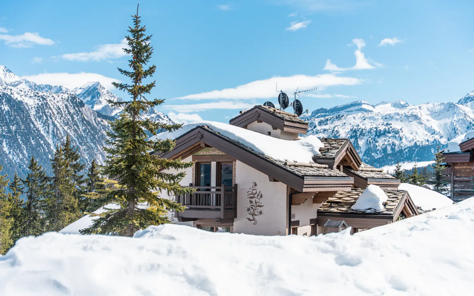 Chalet cubierto de nieve y rodeado de pinos, con majestuosas montañas nevadas de fondo bajo un cielo azul despejado.