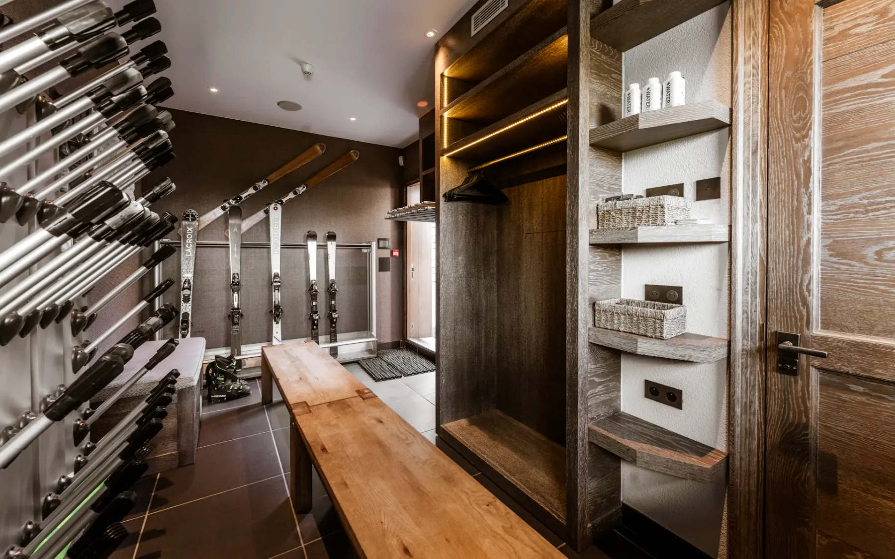 Cuarto para guardar material de esquí con bancos de madera, estantes, bastones de esquí, esquís y artículos de aseo ordenados en los estantes.