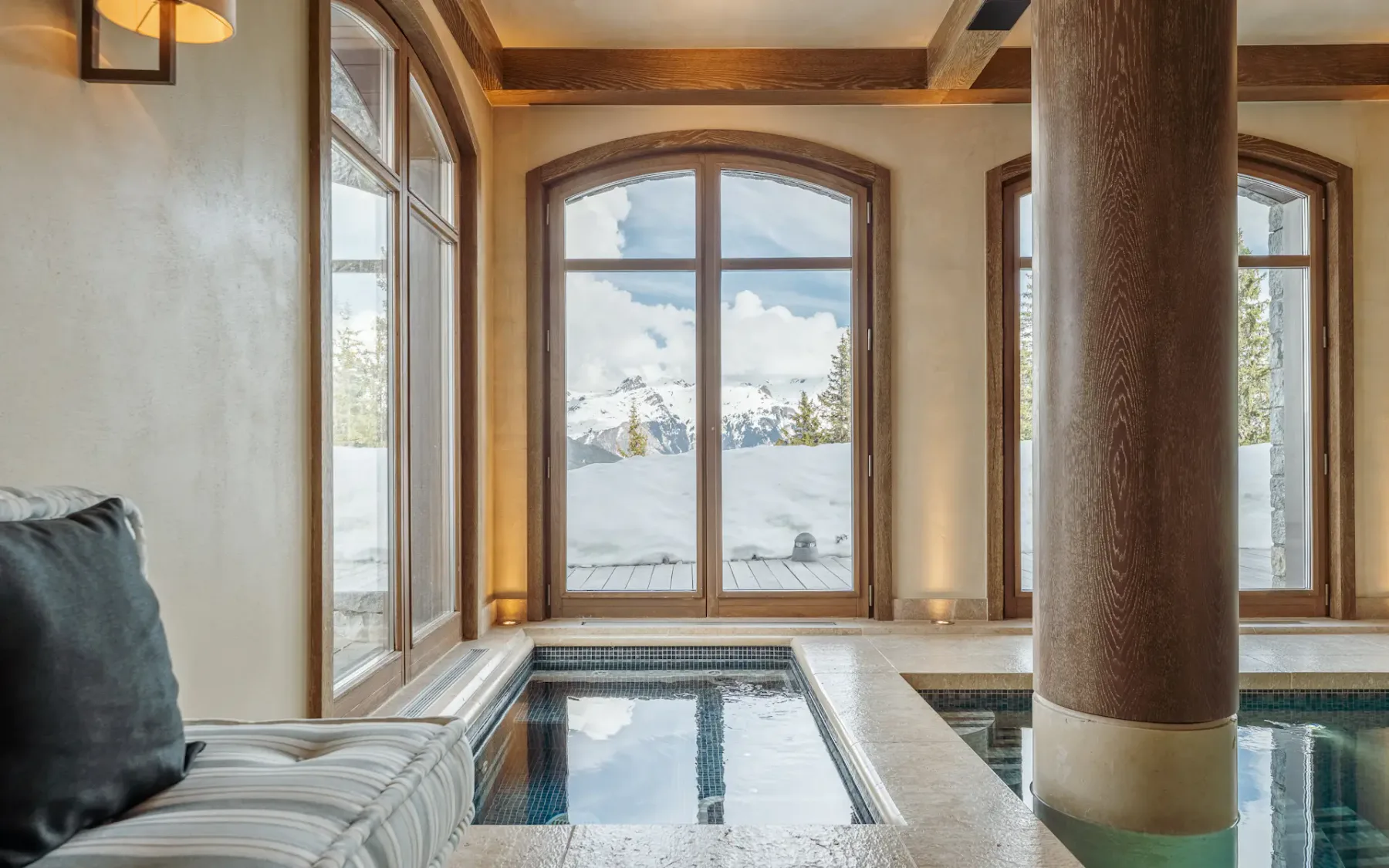 Piscina cubierta bajo vigas de madera, con ventanas arqueadas que dan a un paisaje nevado.