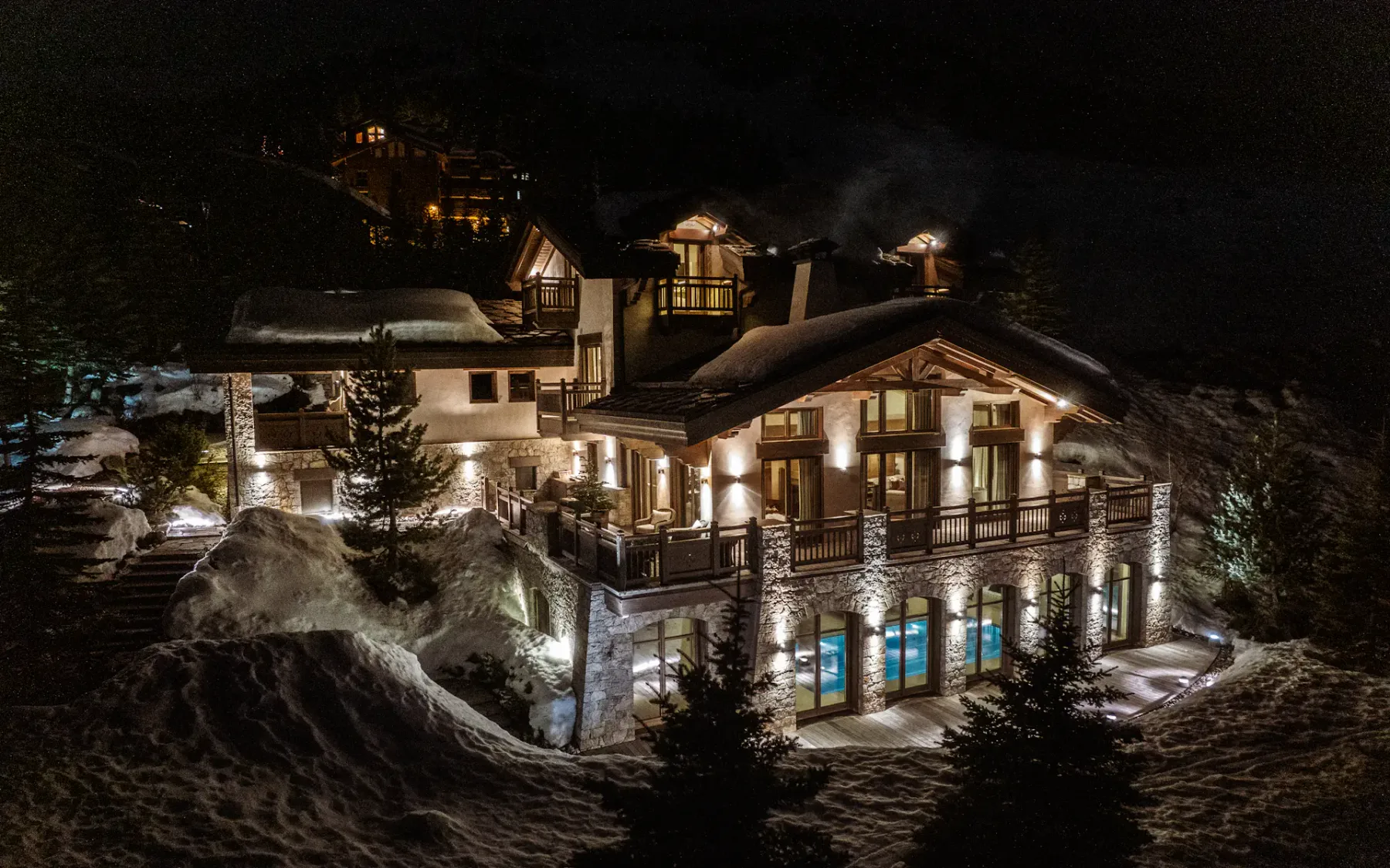 Chalet lujoso e iluminado en un paisaje nevado por la noche, con grandes ventanales, detalles de madera y pinos alrededor.