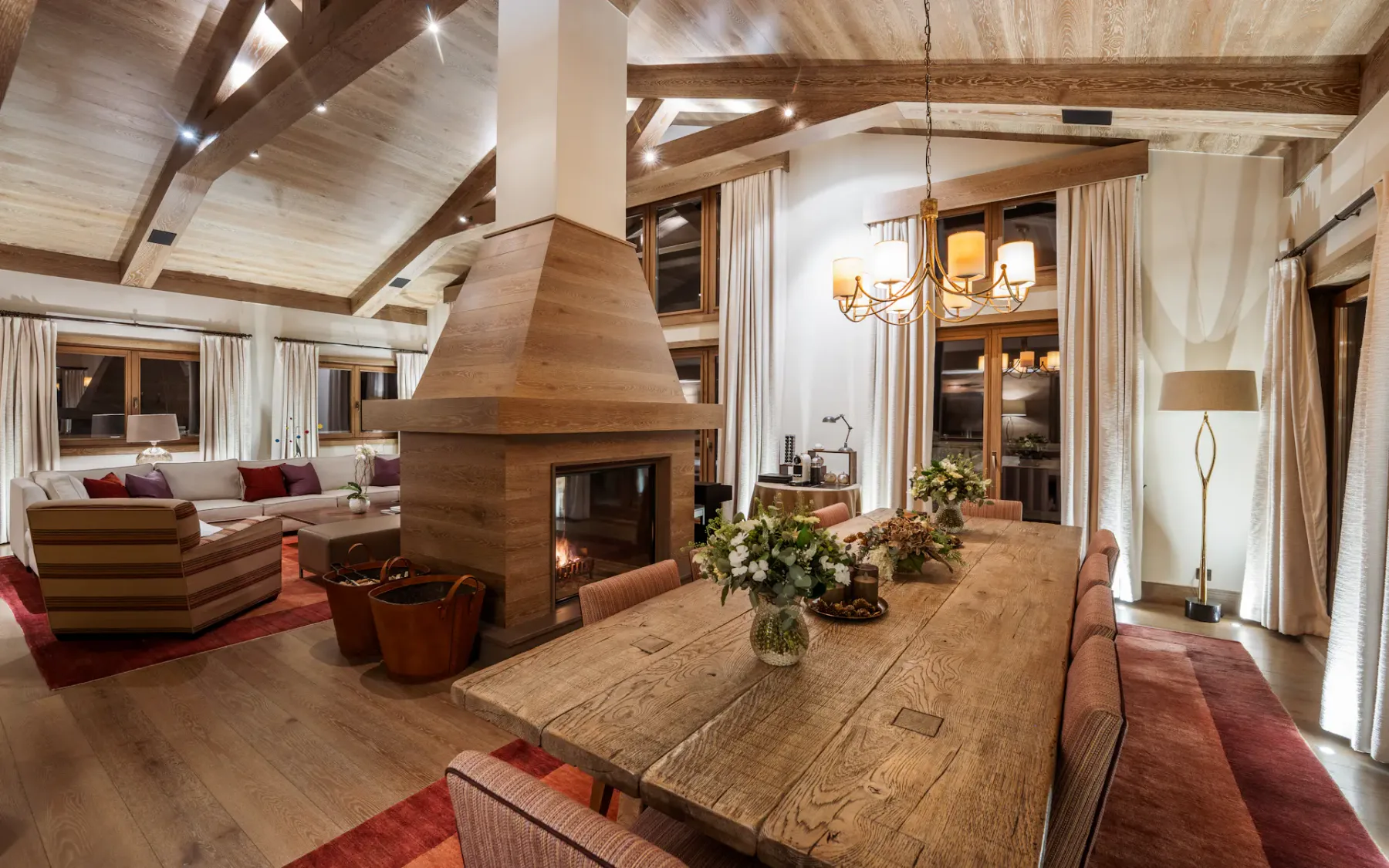 Acogedora zona de estar y comedor con vigas de madera, una chimenea central, una mesa larga, sillas acolchadas e iluminación cálida.