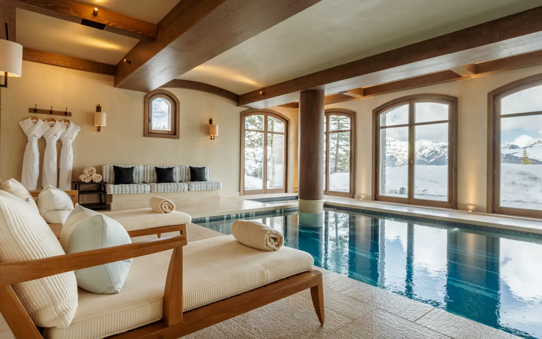Una piscina cubierta junto a asientos acolchados, con grandes ventanas que dan a montañas nevadas.