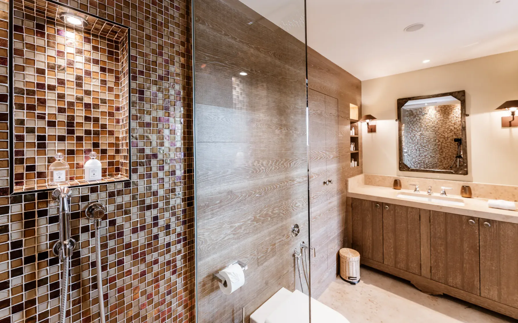 Baño moderno con ducha revestida de mosaico, puerta de cristal, mueble de madera con dos lavabos, gran espejo e iluminación cálida.