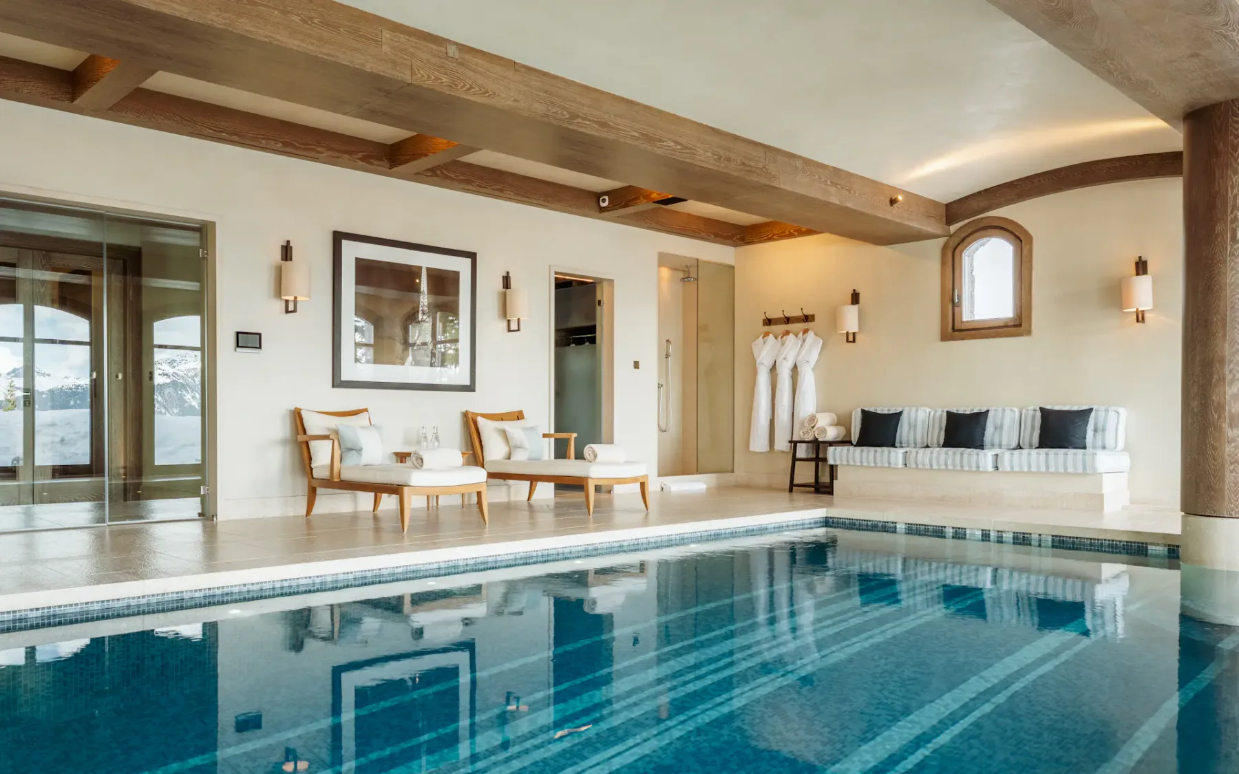 Lujosa zona de piscina cubierta con vigas de madera, tumbonas, un sofá y albornoces colgados junto a la pared, que transmite un ambiente sereno y elegante.
