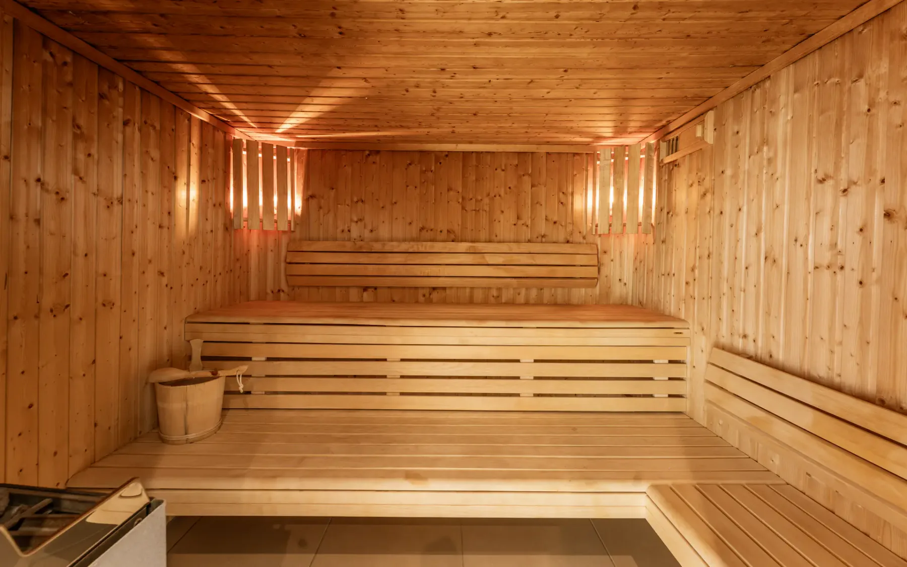 Interior de sauna de madera con iluminación cálida, bancos a dos alturas, un cubo con cucharón y un calentador en la esquina.