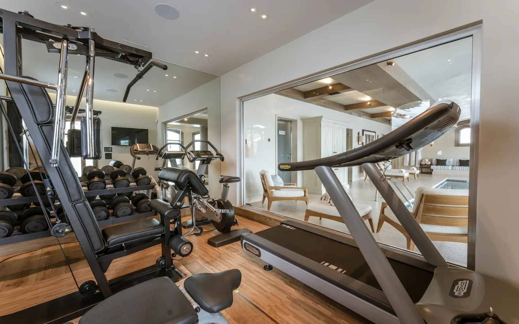 Gimnasio doméstico moderno con cinta de correr, bicicleta estática y pesas. Gran espejo y pared de cristal con vistas a una piscina y una zona de asientos.