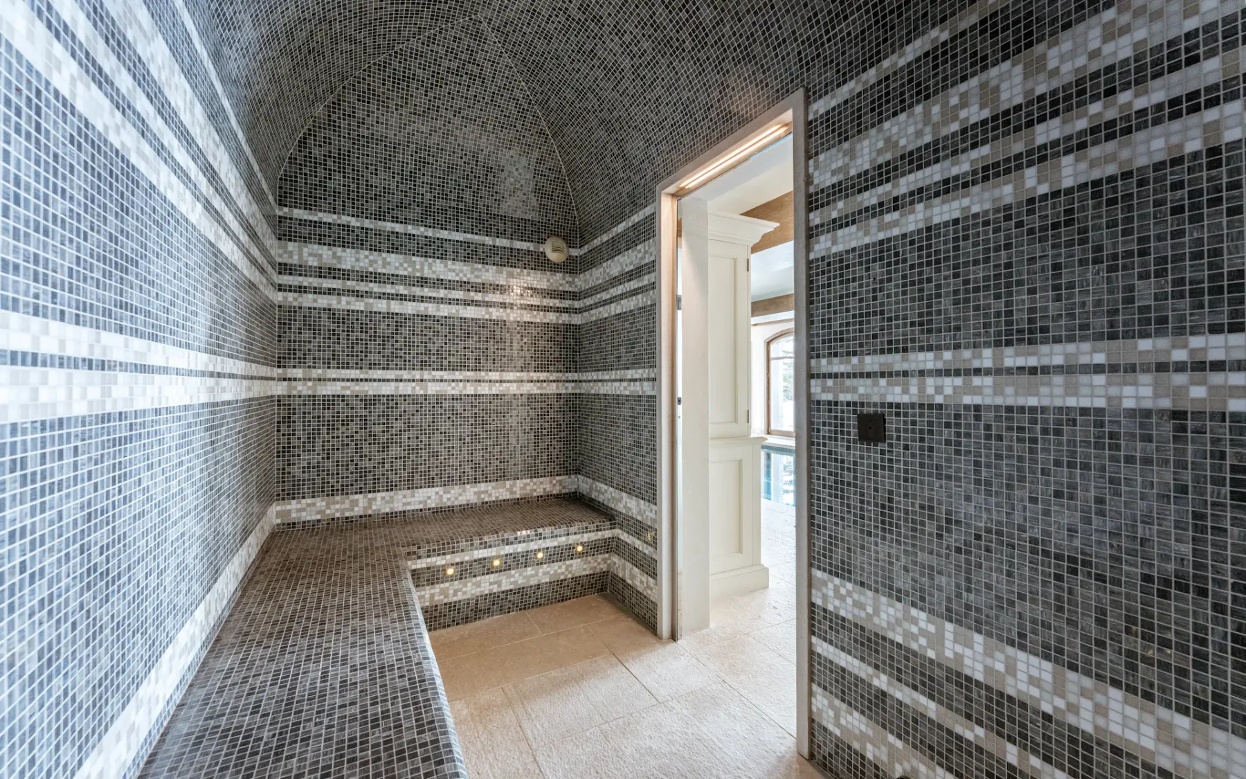 Baño de vapor revestido de mosaico, con techo abovedado y banco integrado, y una puerta parcialmente abierta que da a una zona contigua luminosa.