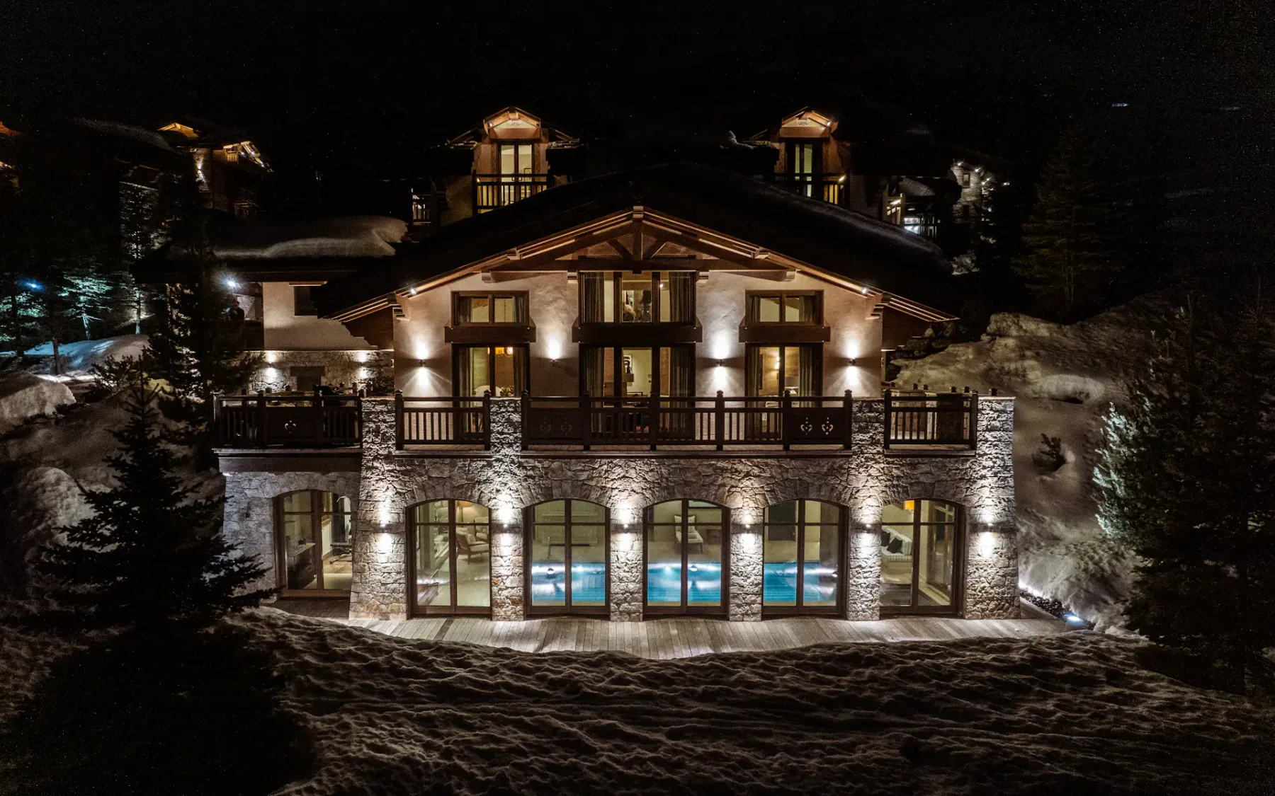 Chalet de montaña iluminado por la noche, con grandes ventanales y un paisaje nevado, rodeado de árboles.