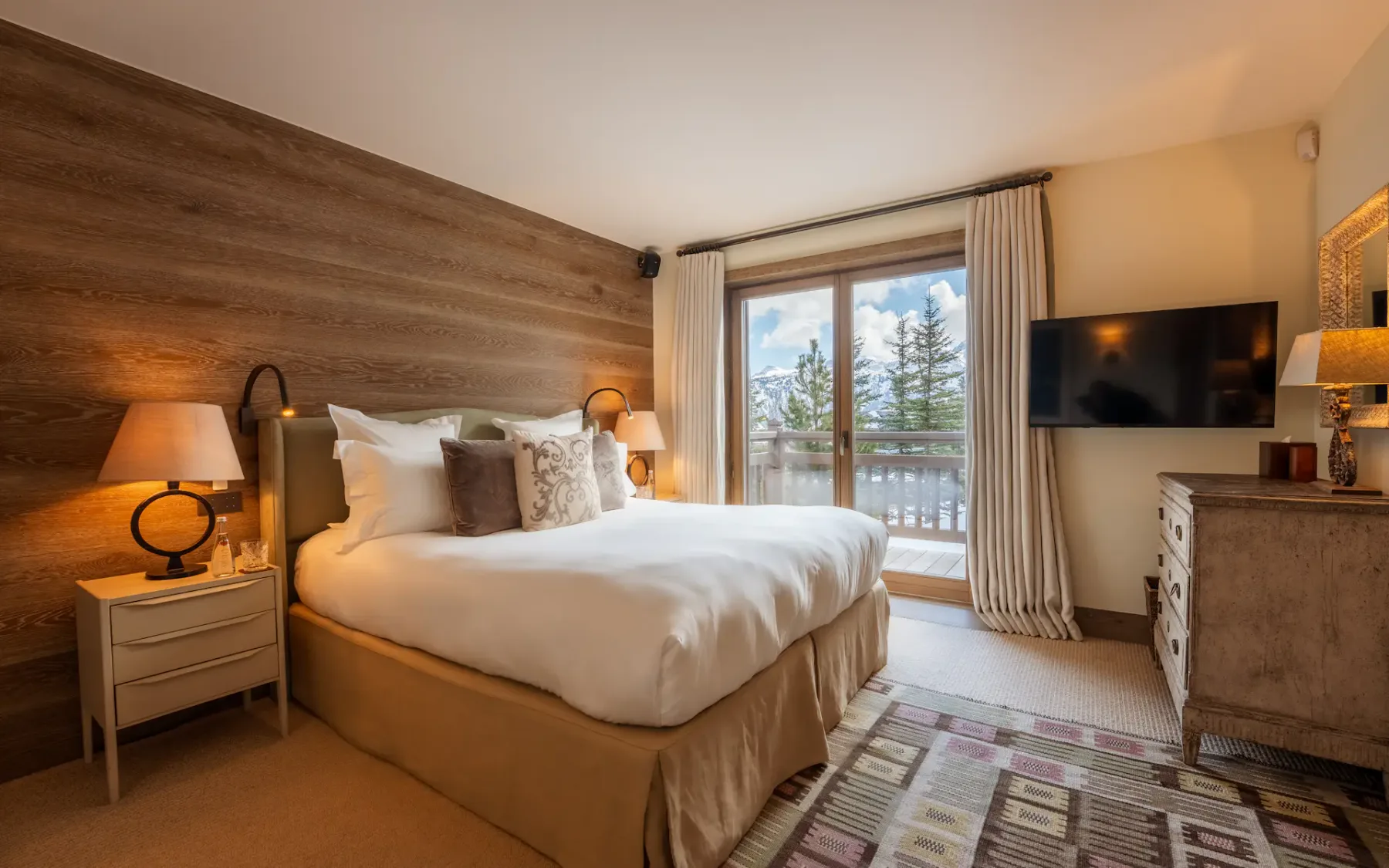Acogedor dormitorio con una cama grande, una pared decorativa de madera, televisor y una ventana con cortinas que da a un balcón con árboles en el exterior.