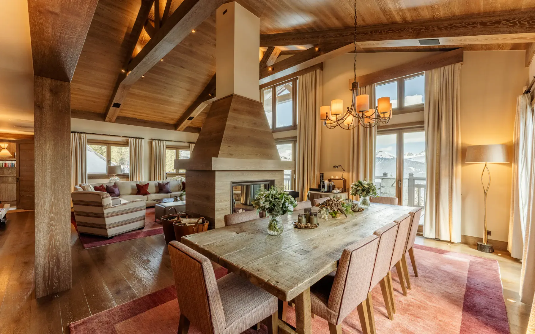 Acogedora zona de estar y comedor con techo de madera, grandes ventanas, una chimenea central y una mesa preparada para ocho personas, con vistas a un paisaje pintoresco.