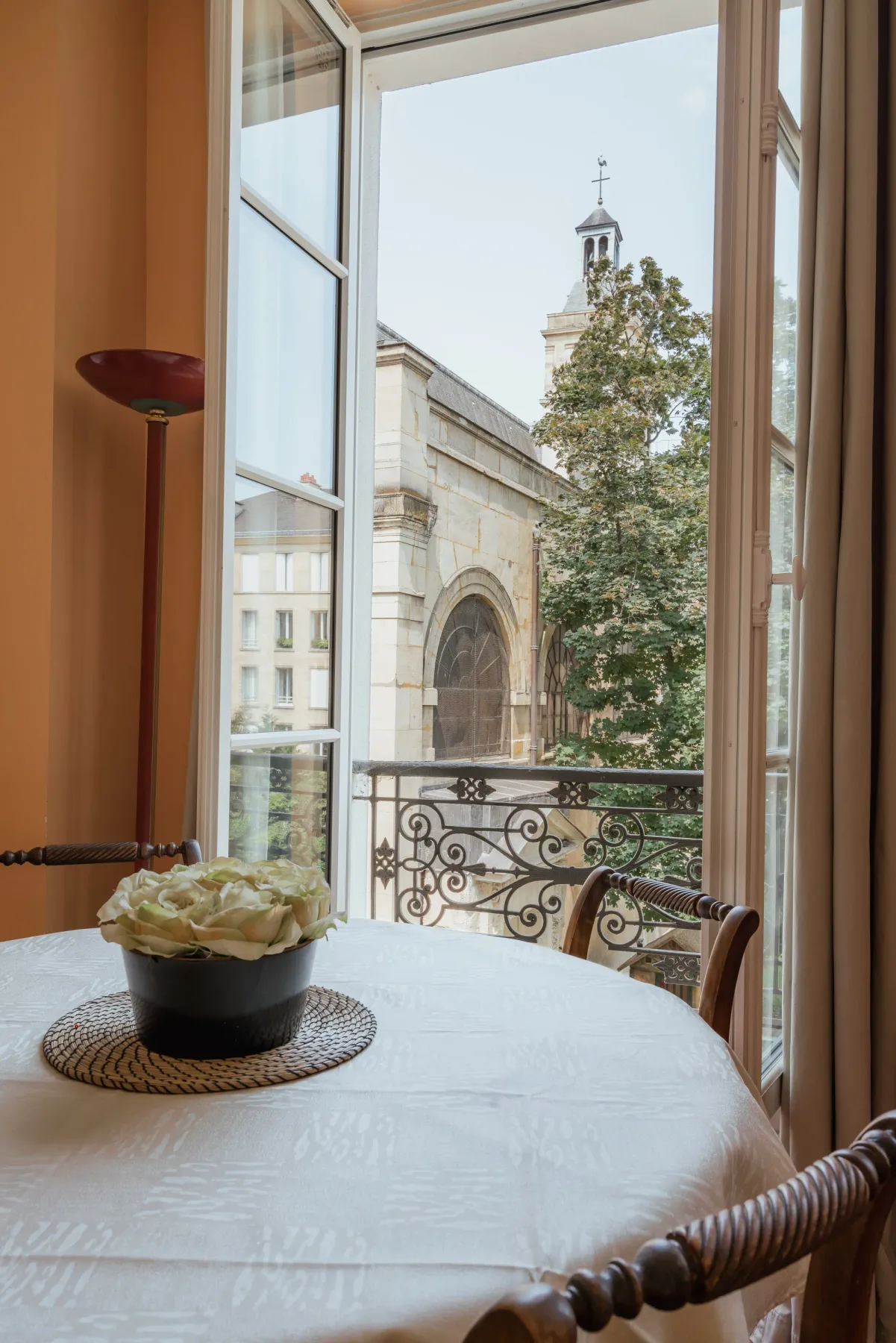 A dining table beside open windows overlooks a stone arch and leafy tree.