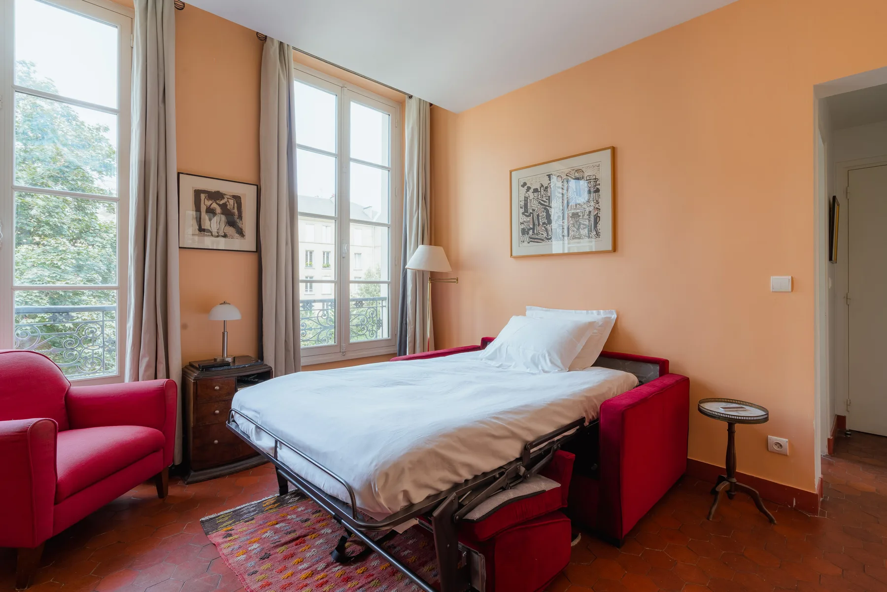 A bedroom with a bed, red seating, and tall windows.