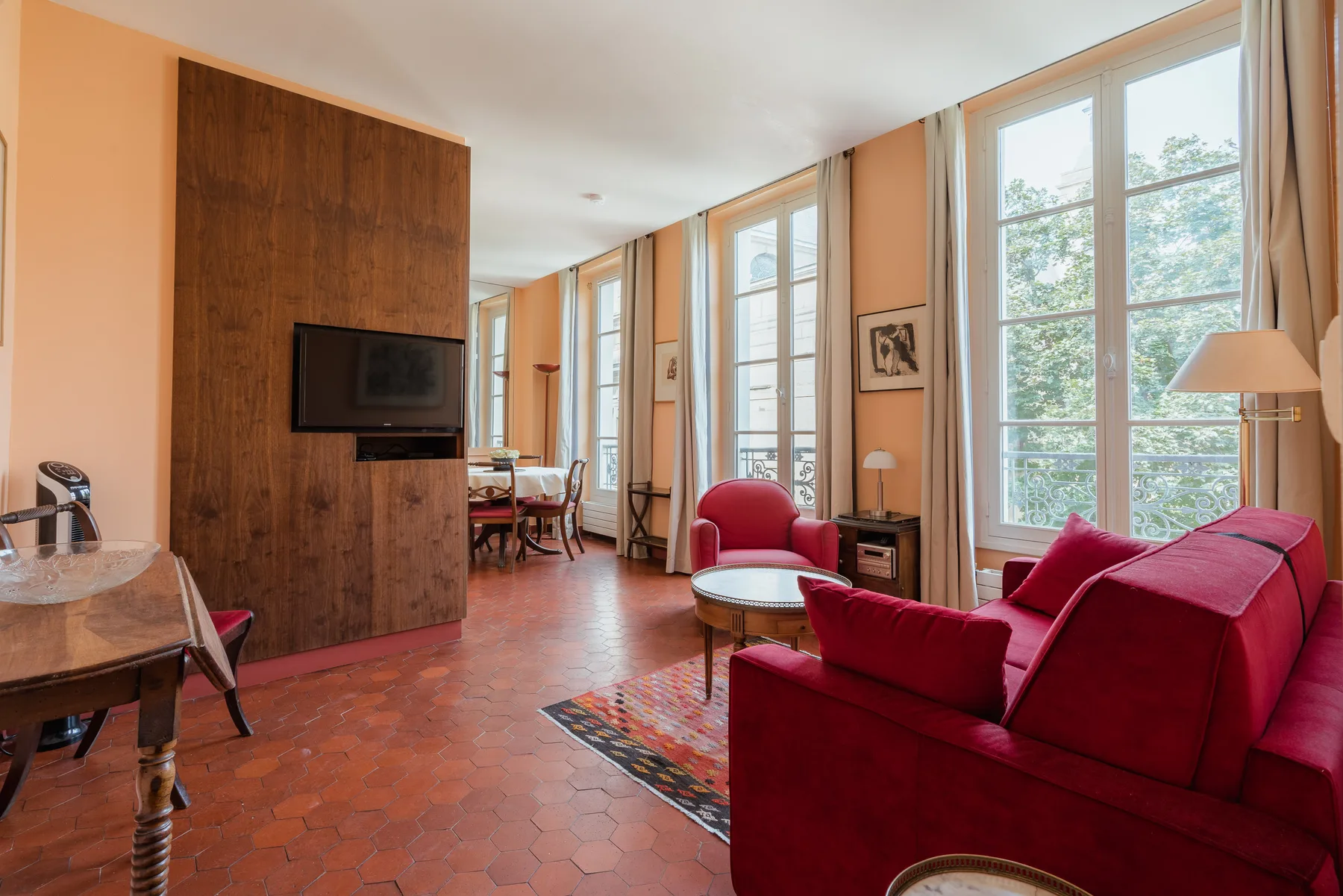 A bright living room has red seating, tall windows, and a wooden television panel.