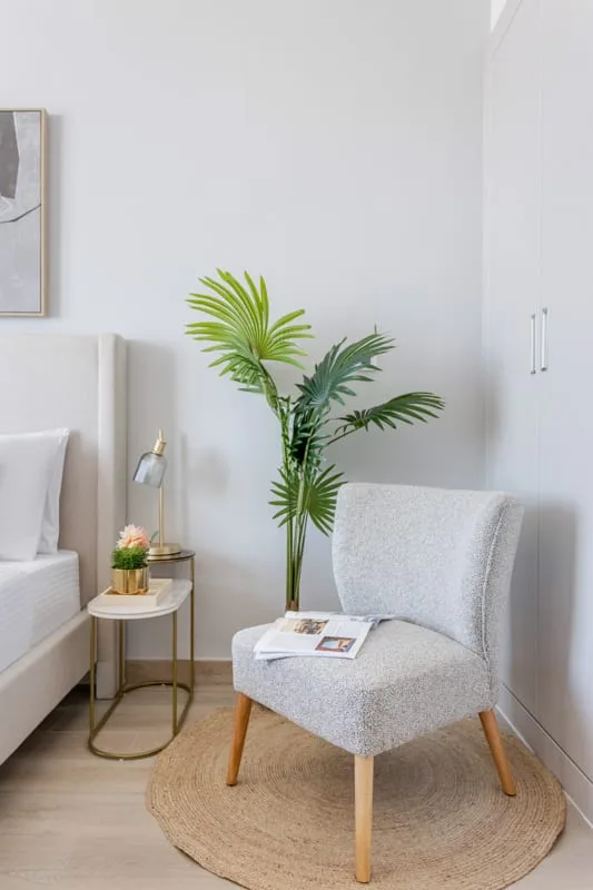 Un rincón luminoso del dormitorio tiene un sillón junto a la cama y una palmera en maceta.
