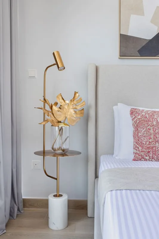 Un dormitorio con una cama, una lámpara dorada y hojas decorativas sobre una mesa auxiliar.