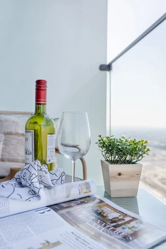 Una mesa junto a una ventana grande tiene una botella de vino, una copa, una planta en maceta y folletos.