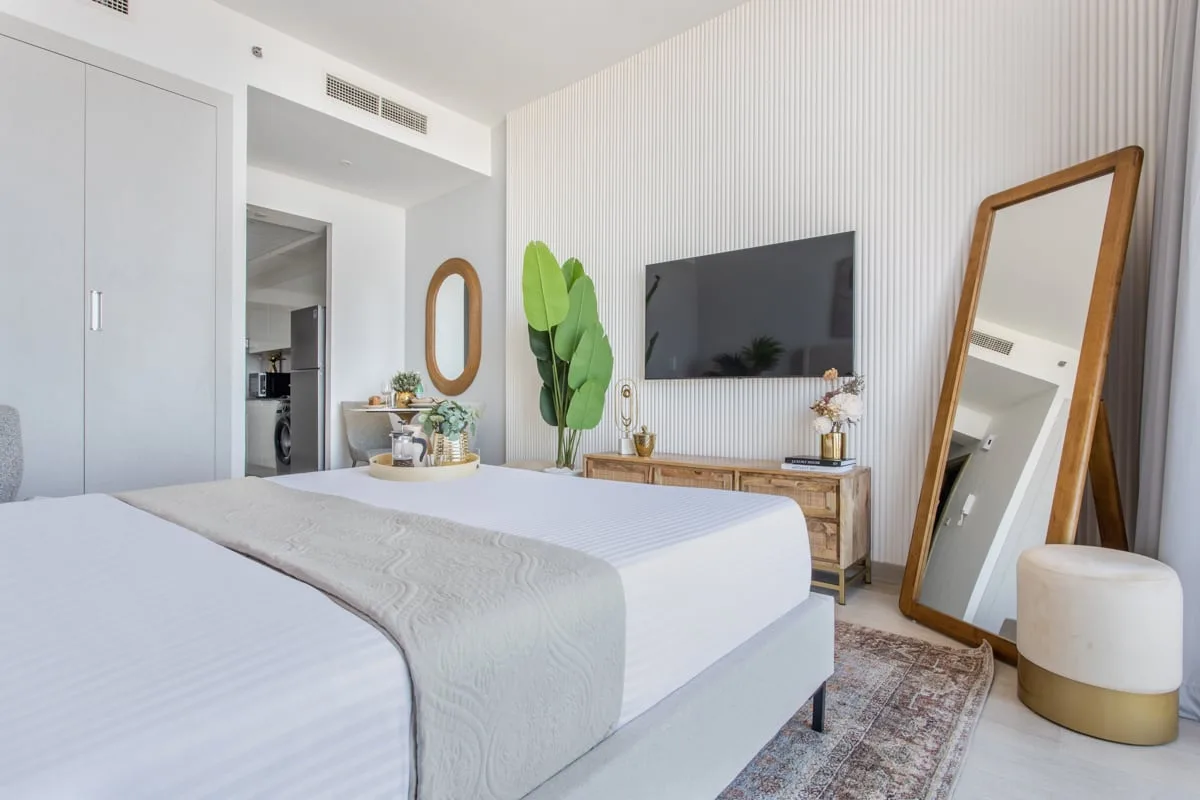 Un dormitorio luminoso con una cama bien tendida, un televisor en la pared y un espejo de cuerpo entero.