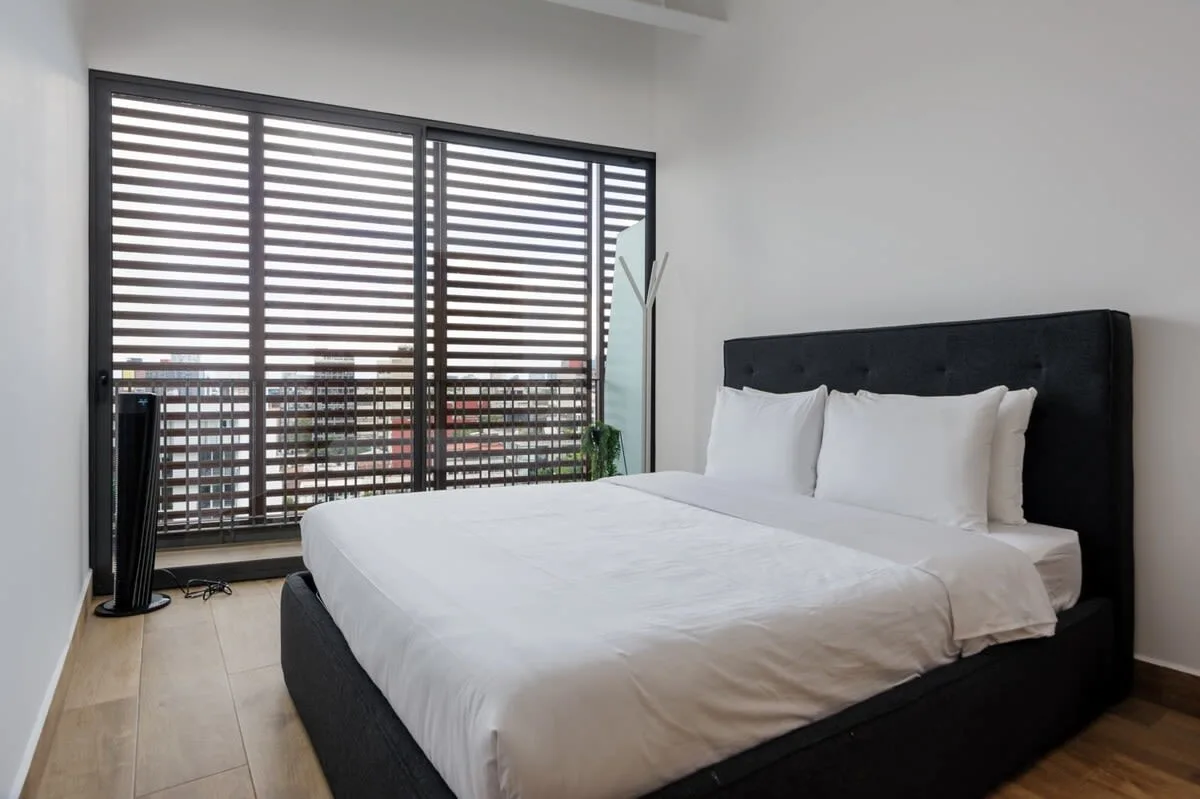 A bedroom with a white bed and dark headboard beside wide windows.