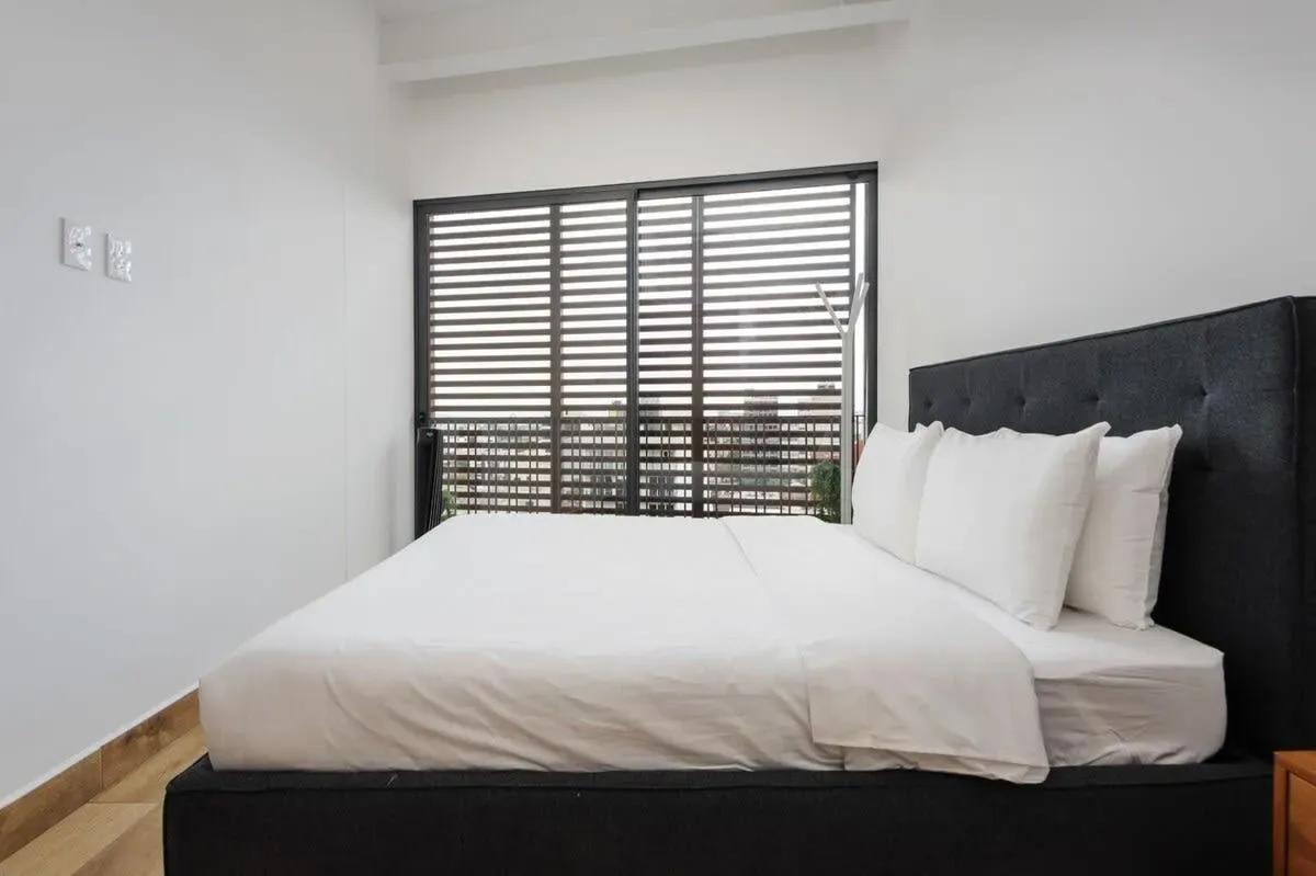 A bedroom with a neatly made bed beneath a wide window with dark blinds.