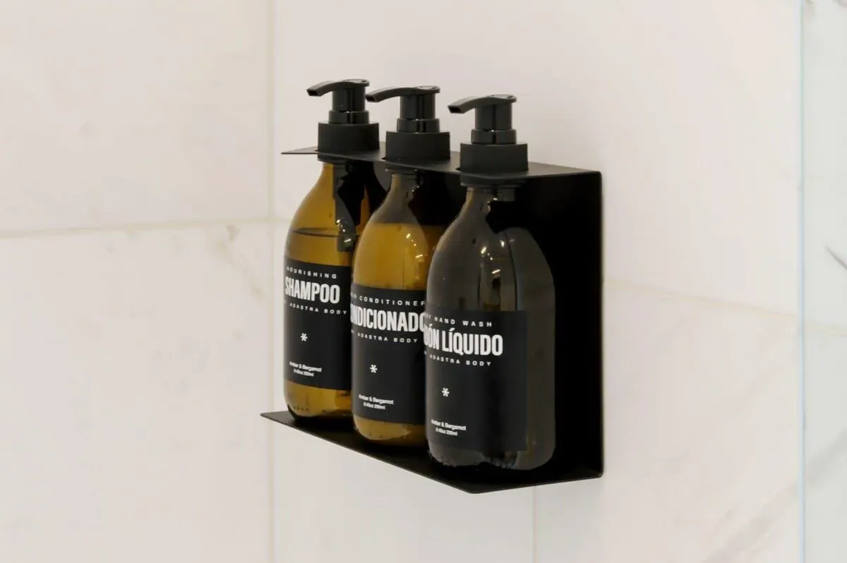 Three pump bottles of shampoo and conditioner sit on a wall-mounted bathroom shelf.