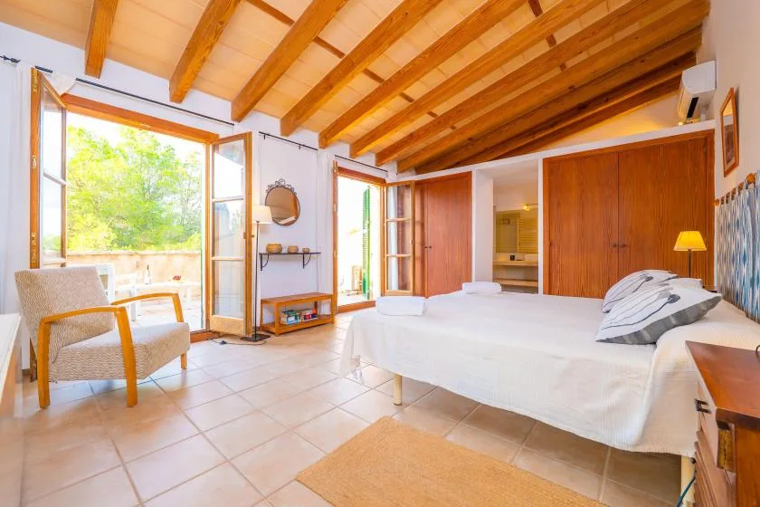 A bright bedroom with a large bed, exposed wooden ceiling beams, and open windows.