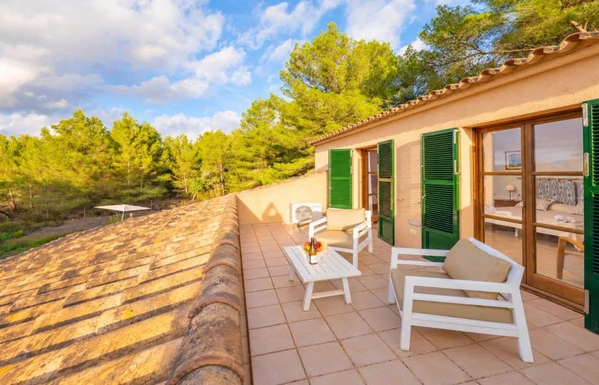 A sunny tiled terrace with outdoor seating beside a house and green shutters.