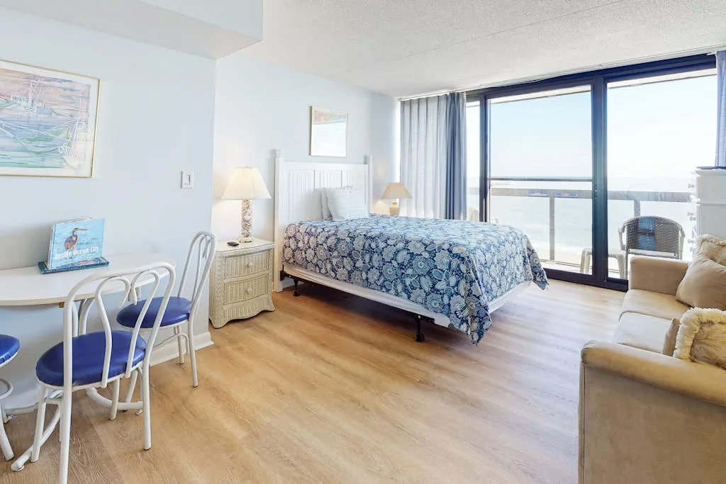 A bright bedroom with a bed, desk, and large windows overlooking the water.