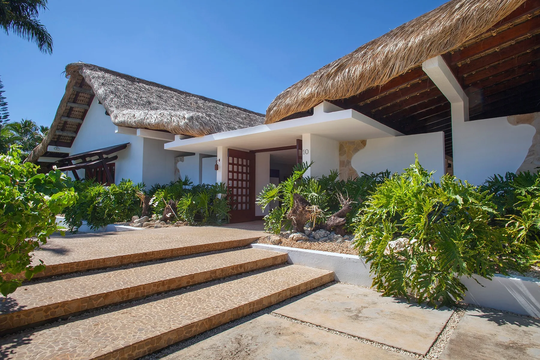 A white building with thatched roof sections, tropical plants, and steps at the entrance.