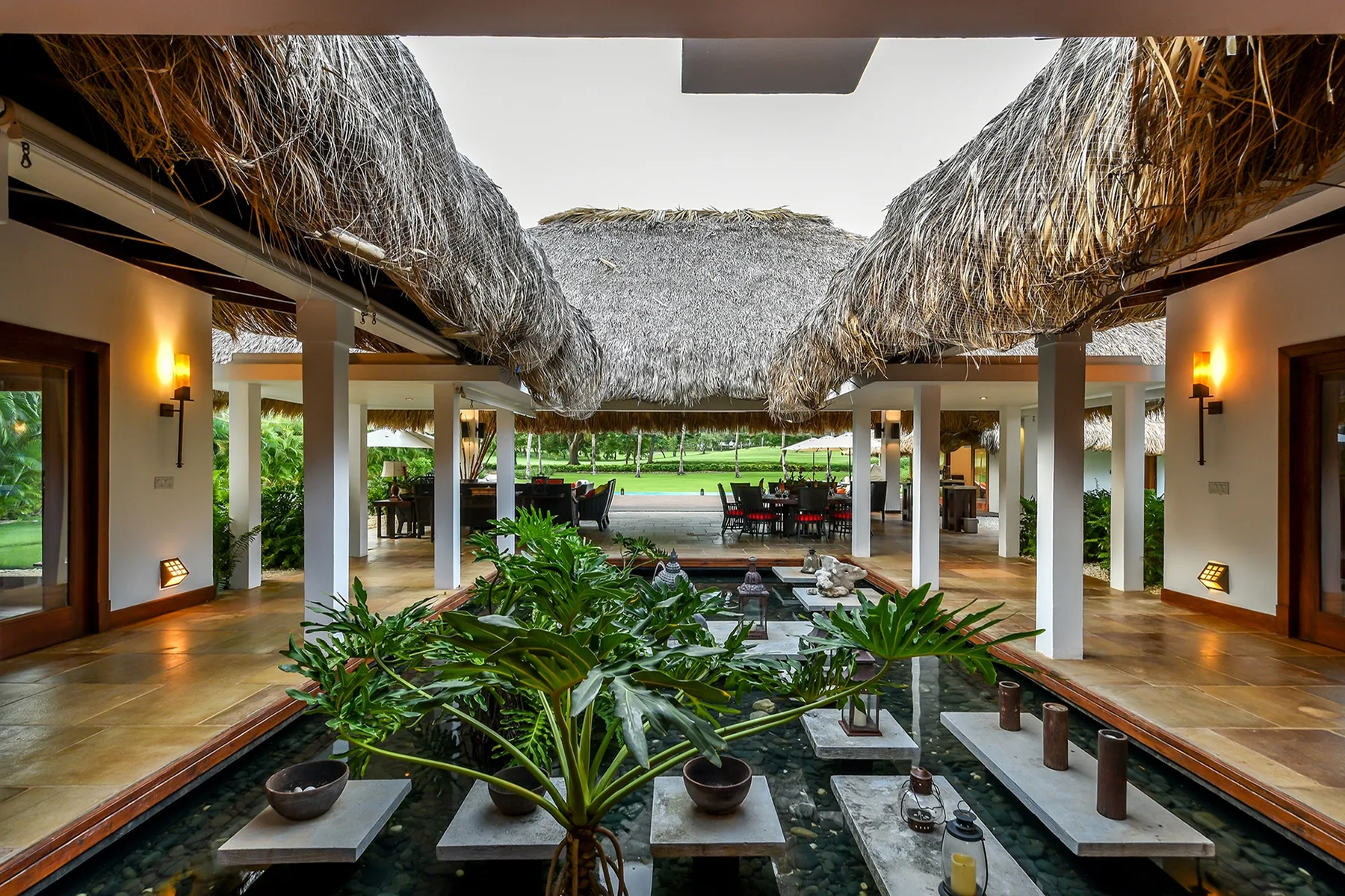 An open-air resort dining area with thatched roofing, tables, and lush plants.