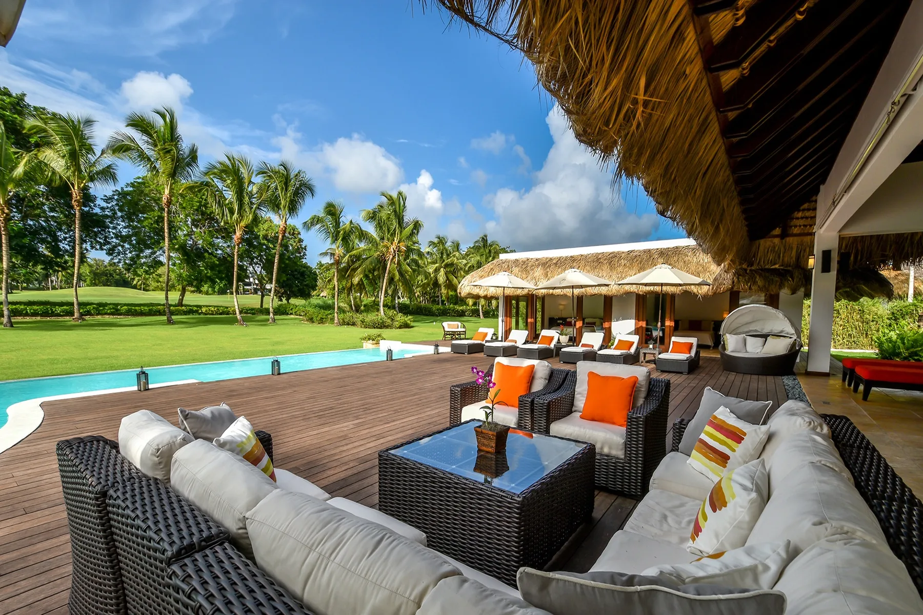 An outdoor lounge with cushioned seating overlooks a swimming pool and palm trees.
