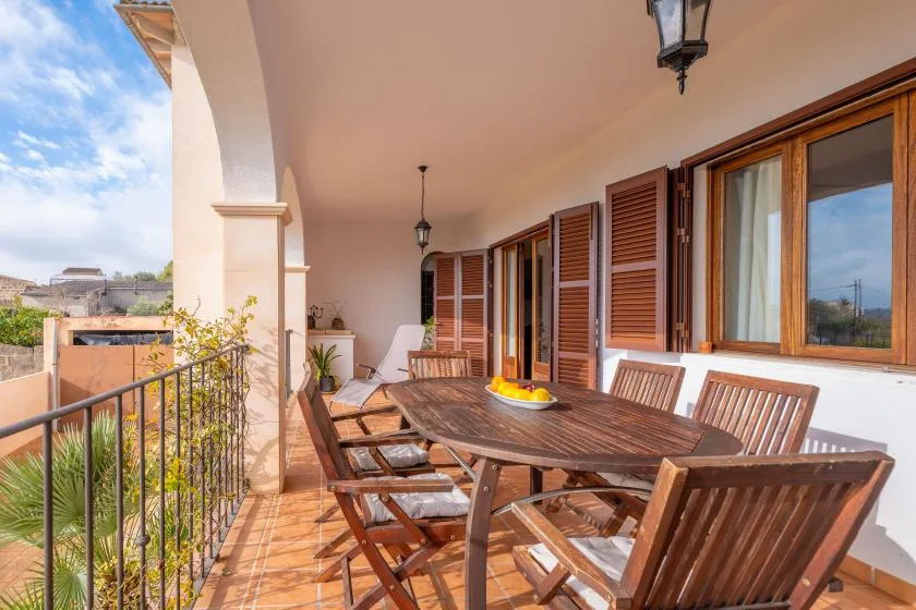 A covered terrace with a wooden dining table and chairs beside open windows.