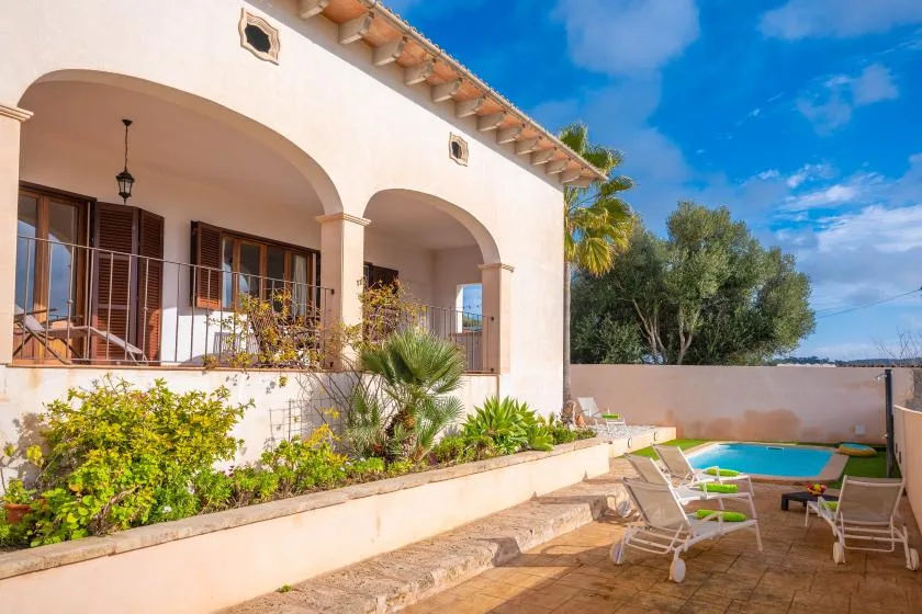 A sunlit house patio overlooks a swimming pool and landscaped garden.