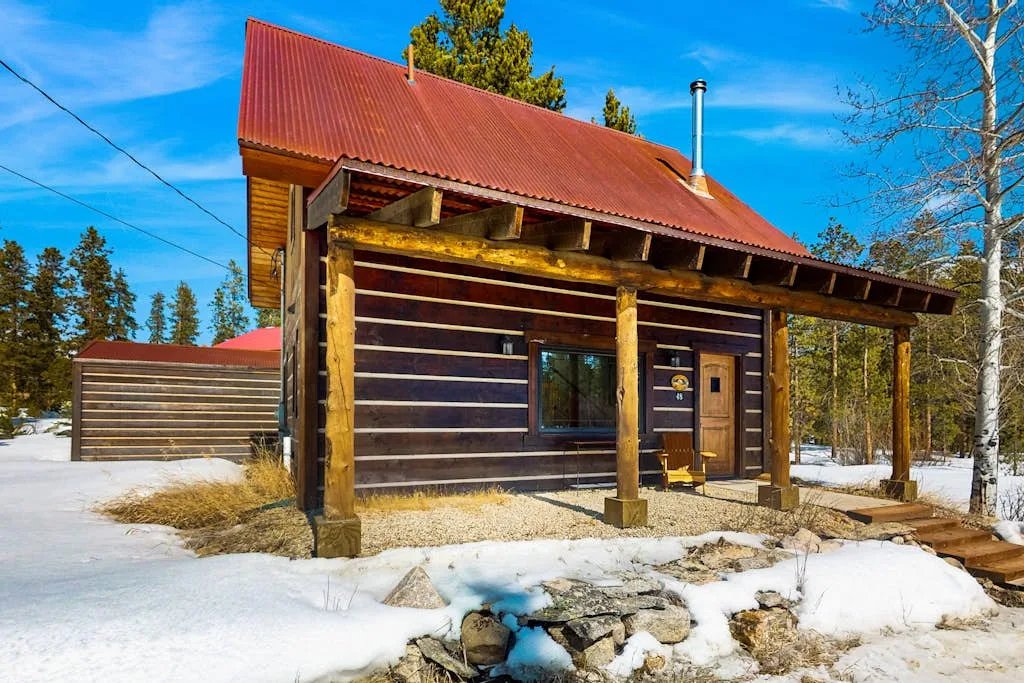 A wooden cabin stands in a snowy clearing beneath a bright blue sky.